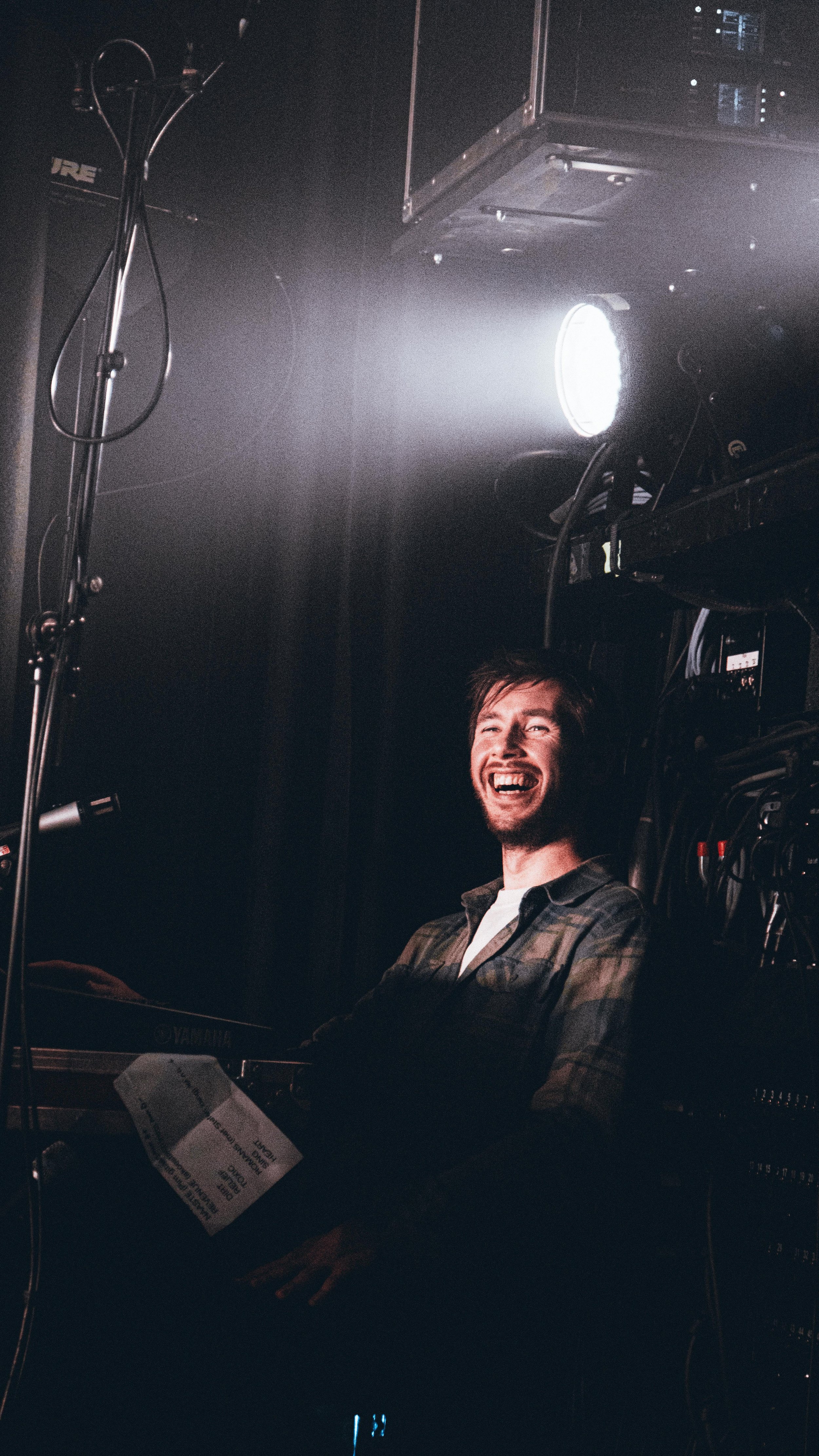 A man with a beard and mustache, wearing a plaid shirt, is laughing while sitting at a piano in a dark setting with bright stage lights shining in the background.