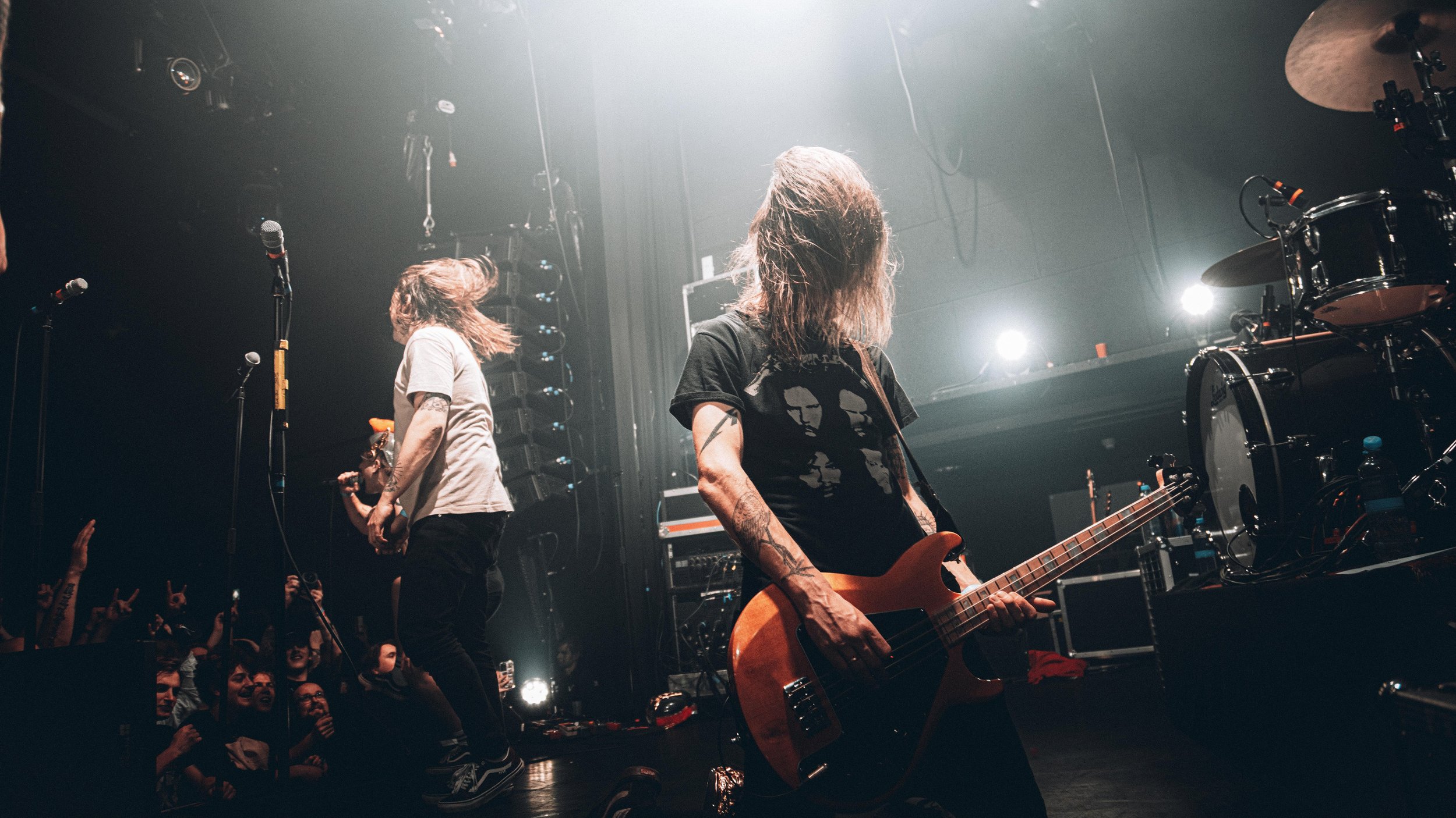 Musicians performing on stage during a concert, with a guitarist in the foreground and a singer in the background, audience cheering