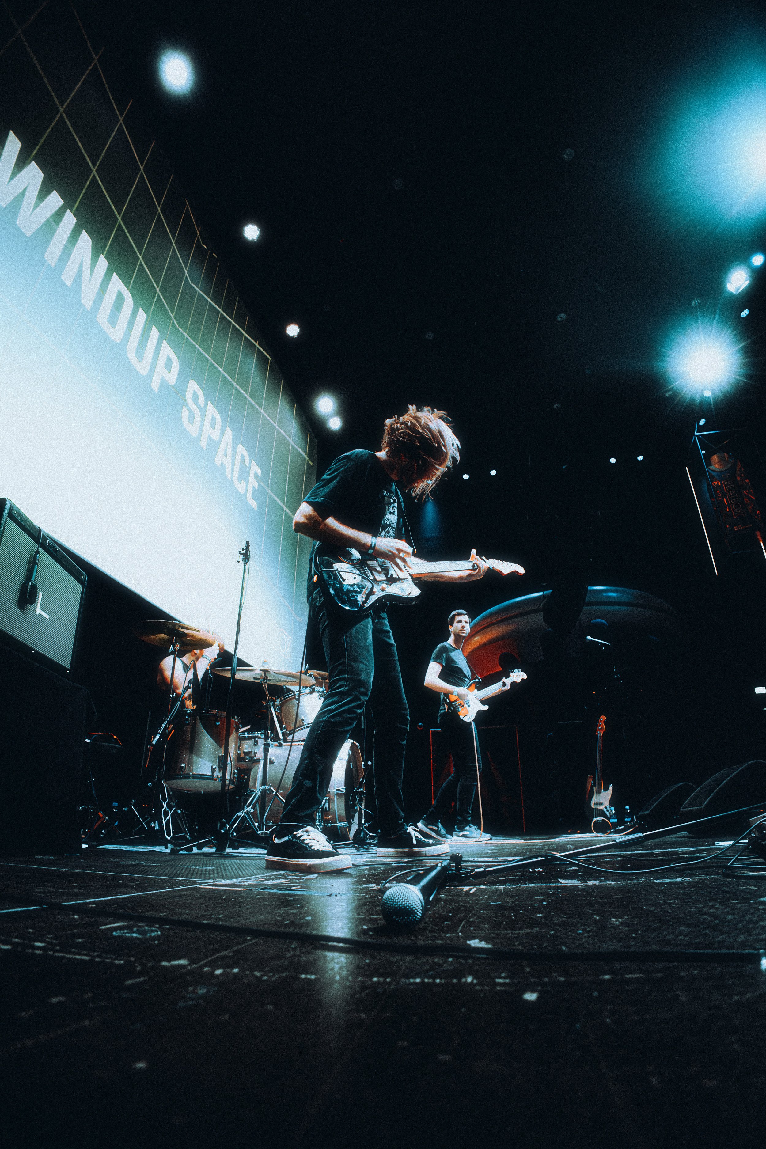 A band performing live on stage with electric guitars, drums, and microphones, with a large screen displaying the words 'WINDUP SPACE' in the background.