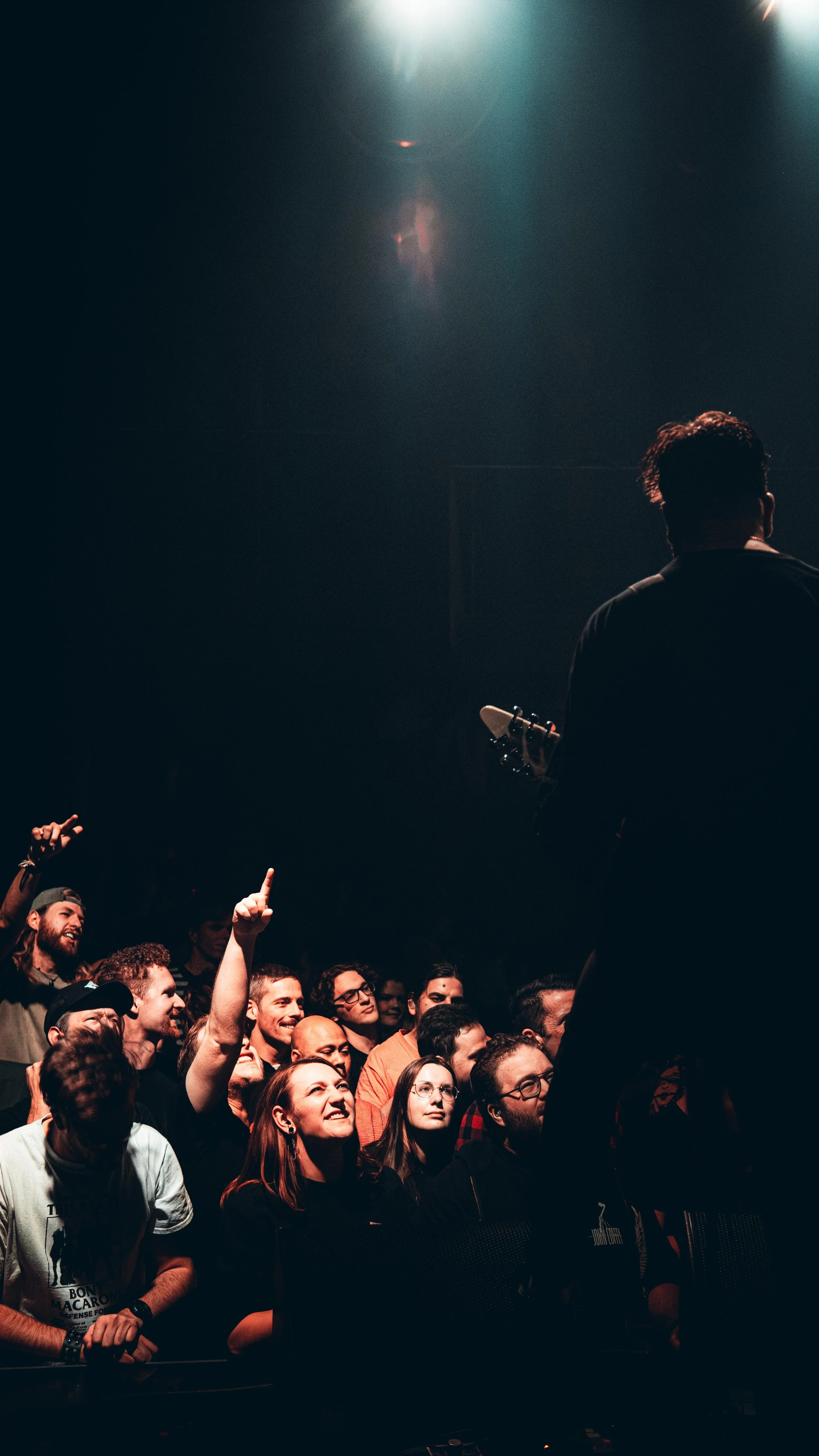 A musician performing on stage with his back to the audience, while the crowd in front of him cheers and some raise their hands.