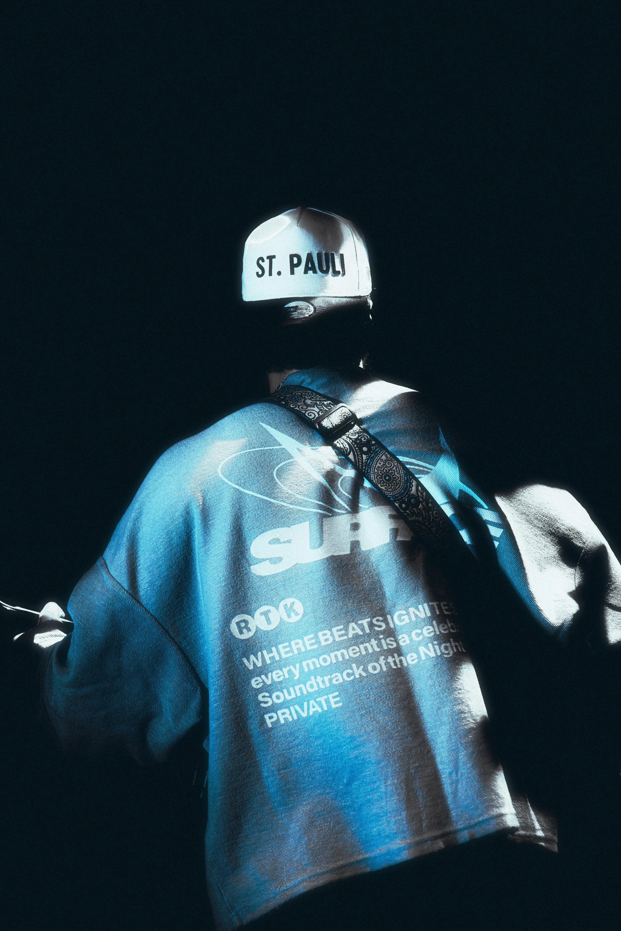 Individual wearing a white helmet with 'ST. PAULI' written on the back, and a blue hoodie with various text and graphics on it, against a dark background.