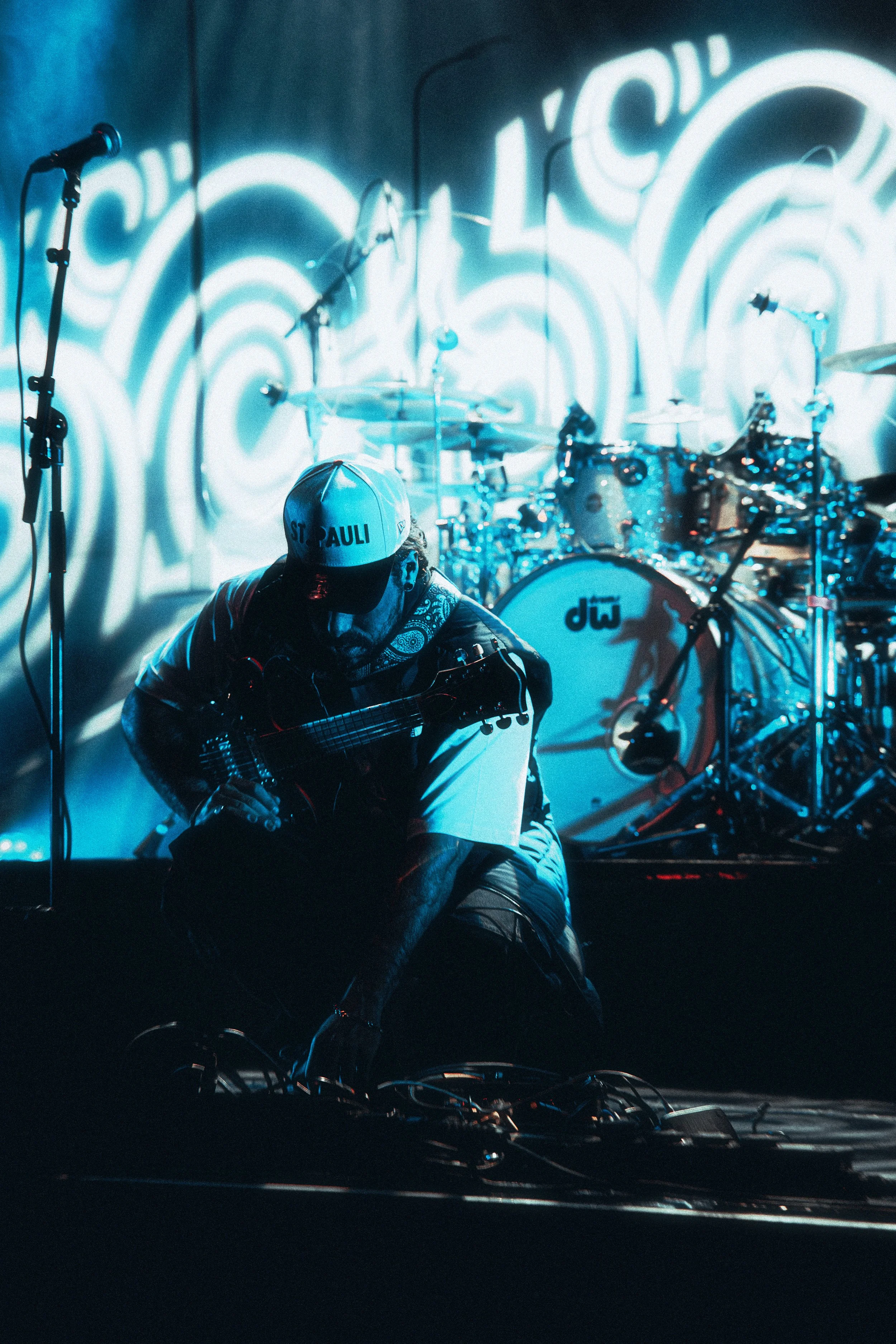 A musician crouched on stage with a guitar, wearing a white cap with 'ST PAULI' written on it, illuminated by blue stage lights, with a drum set in the background.