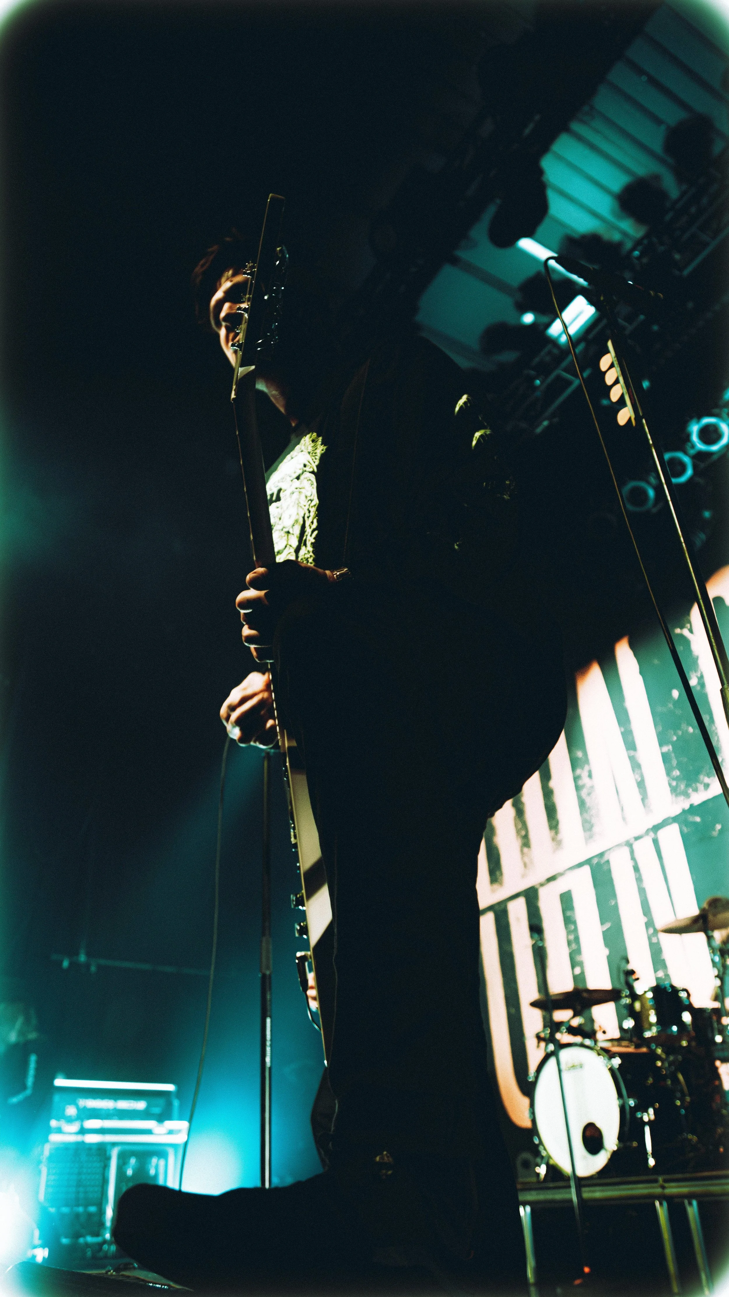 A musician is performing on stage, holding a guitar, with colorful stage lighting and a drum set in the background.