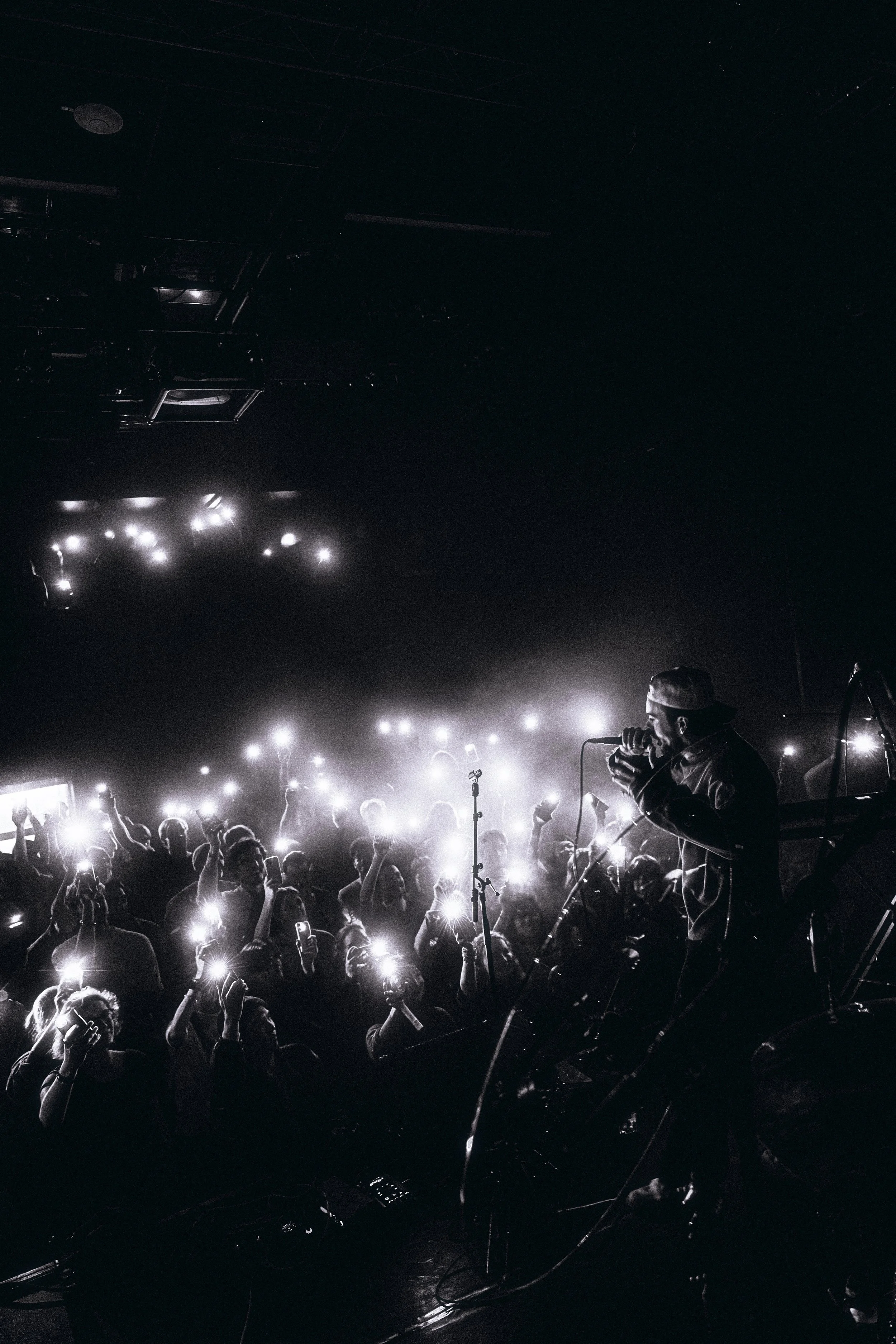 A black-and-white photo of a singer performing on stage at a concert, with bright lights and a crowd of people holding up their phones.