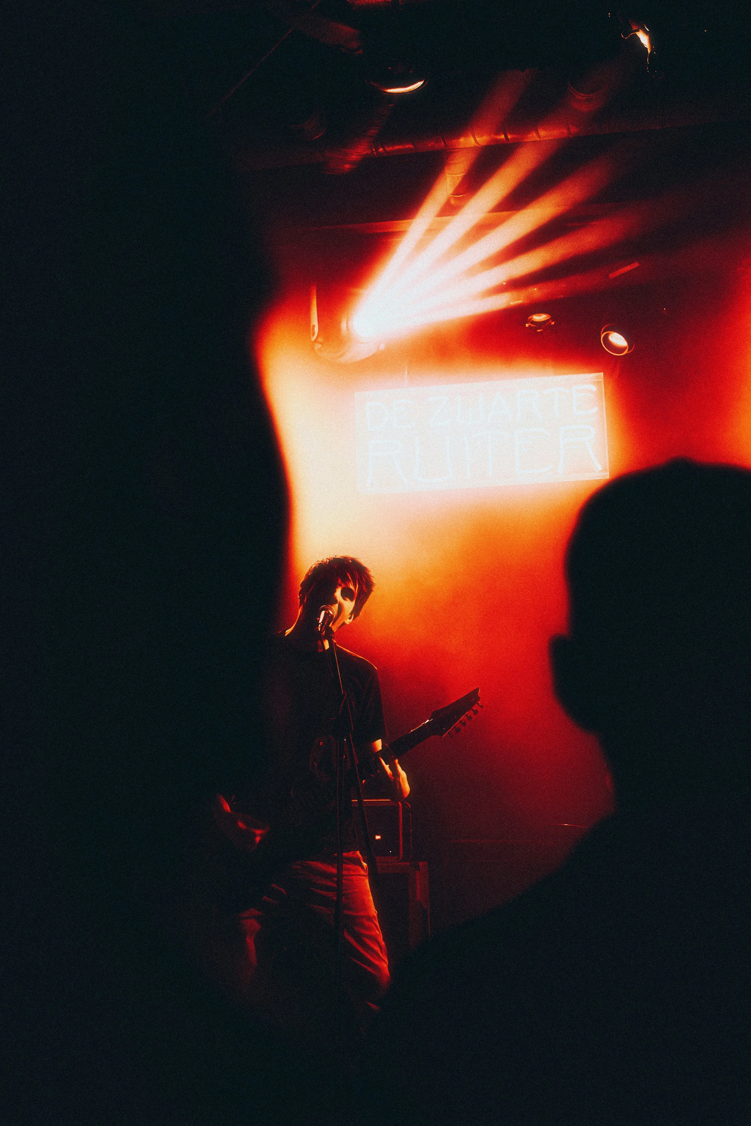 A musician performing on stage with a guitar, illuminated by red stage lights, with a neon sign in the background that reads 'DE ZWARTE RUITER'.