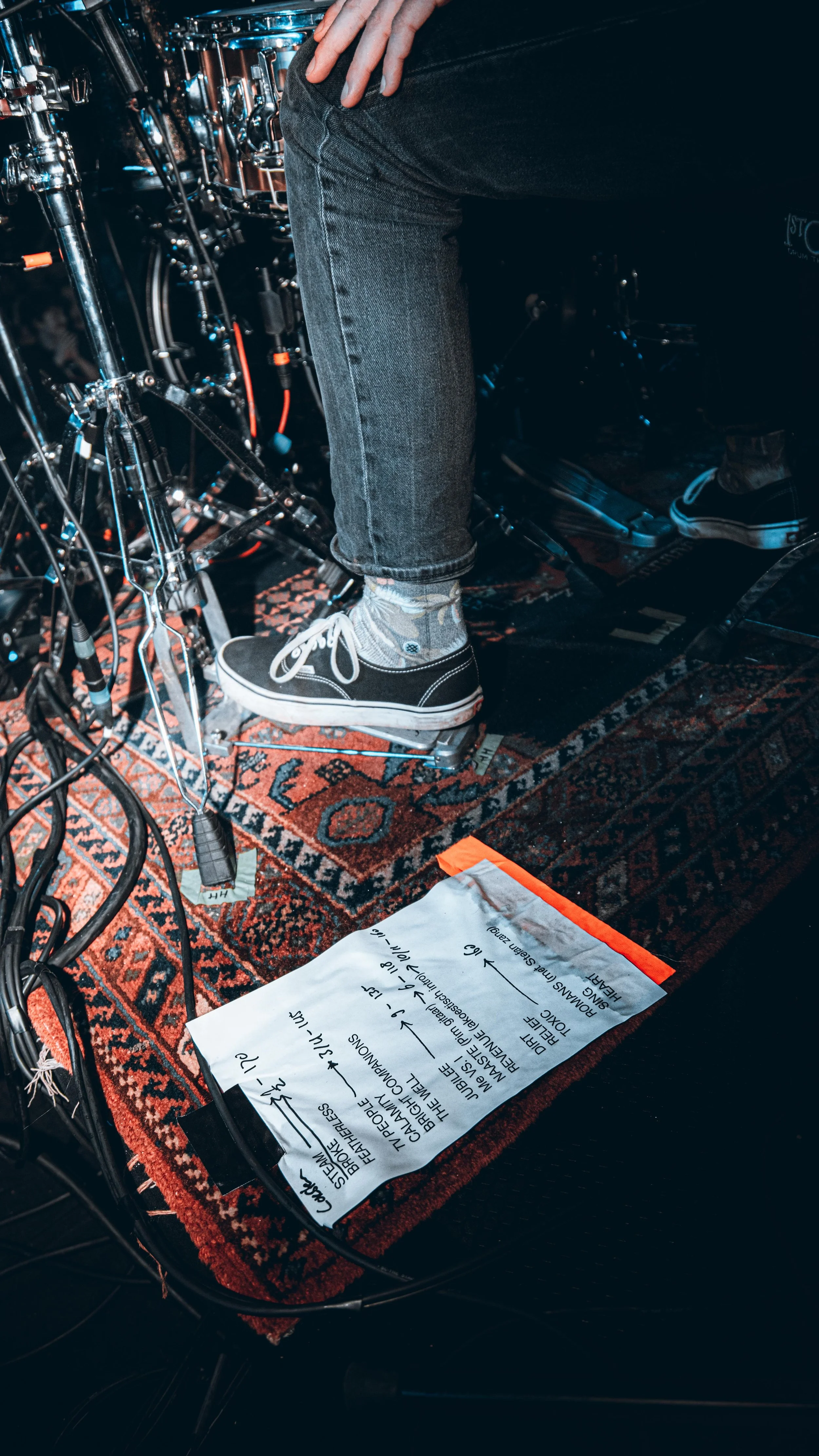 Close-up of a person's lower body and feet on a drum pedal, surrounded by drum equipment, with a setlist on the floor.