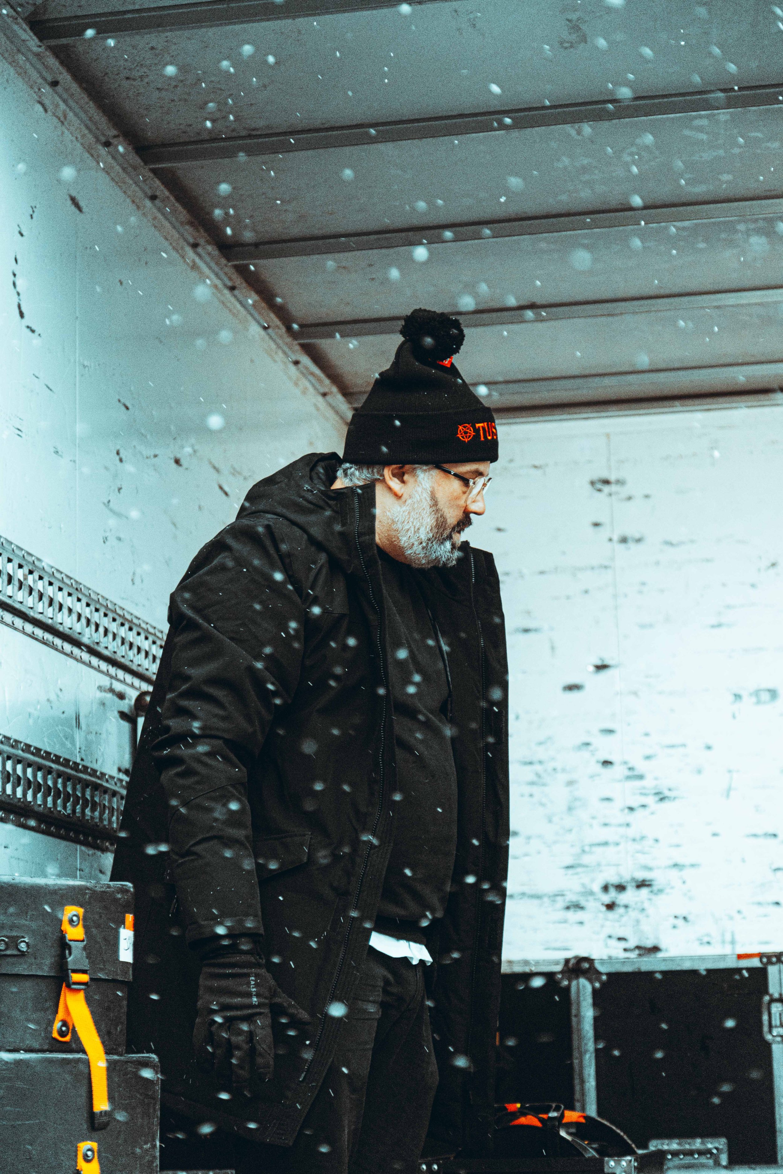 A man with gray hair and beard wearing glasses, a black winter jacket, black gloves, and a black beanie with red text, standing in a snow-covered outdoor area. Snow is falling around him, and he appears to be looking down, possibly at equipment nearb