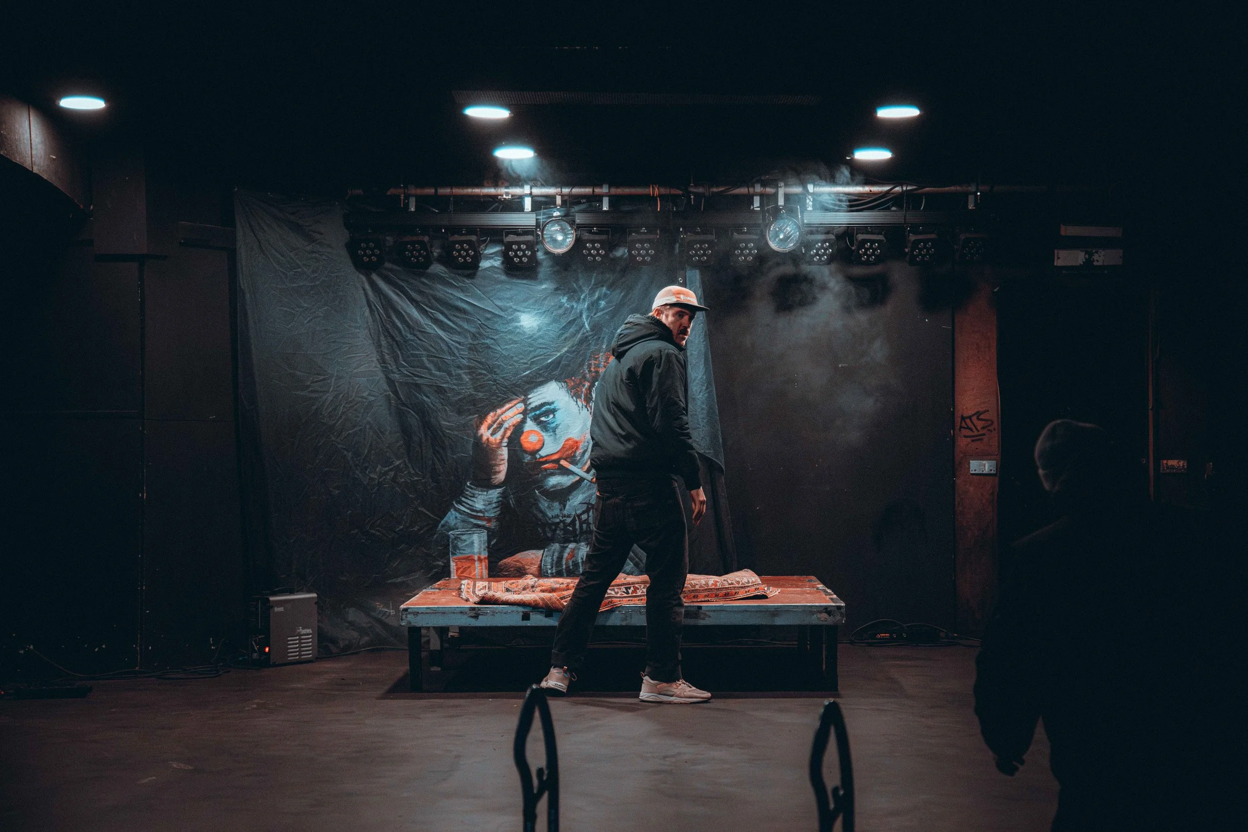 A man in a hoodie and cap standing on a stage with a painted clown backdrop, with stage lights overhead and two people watching.