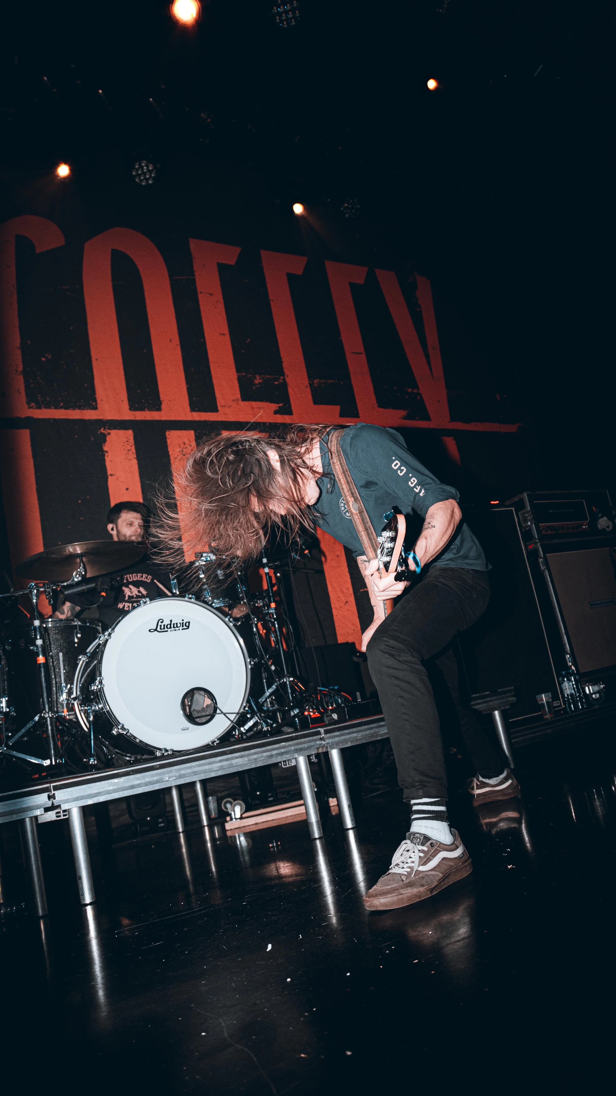 A band performs live on stage. The drummer is playing behind a Ludwig drum set, and the guitarist is bending forward, playing his instrument. There is a large illuminated background with the word 'CLOSED.'