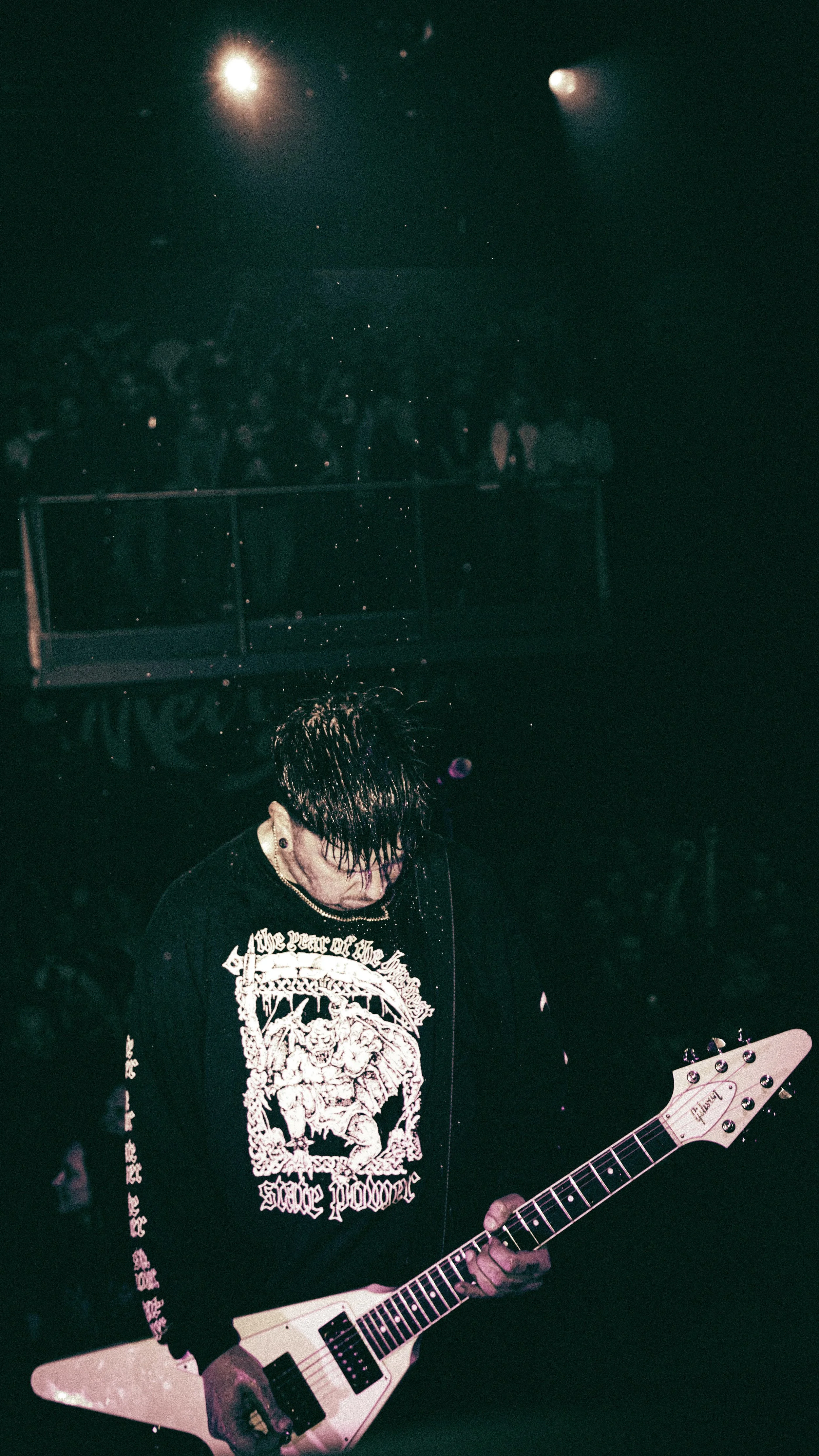 A musician playing a white electric guitar on stage during a concert, with a dark background and an audience visible in the distance.