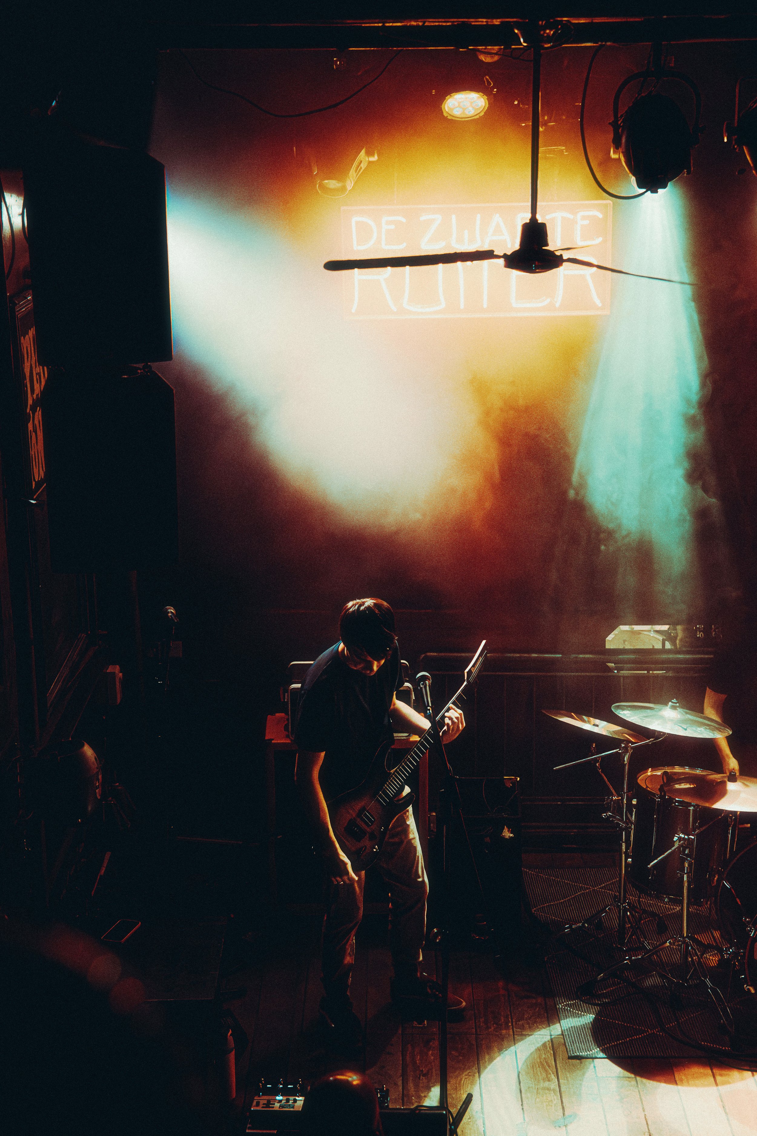 A musician playing an electric guitar on stage, illuminated by colorful stage lights, with a neon sign in the background that reads 'Dezuate Rutter'.