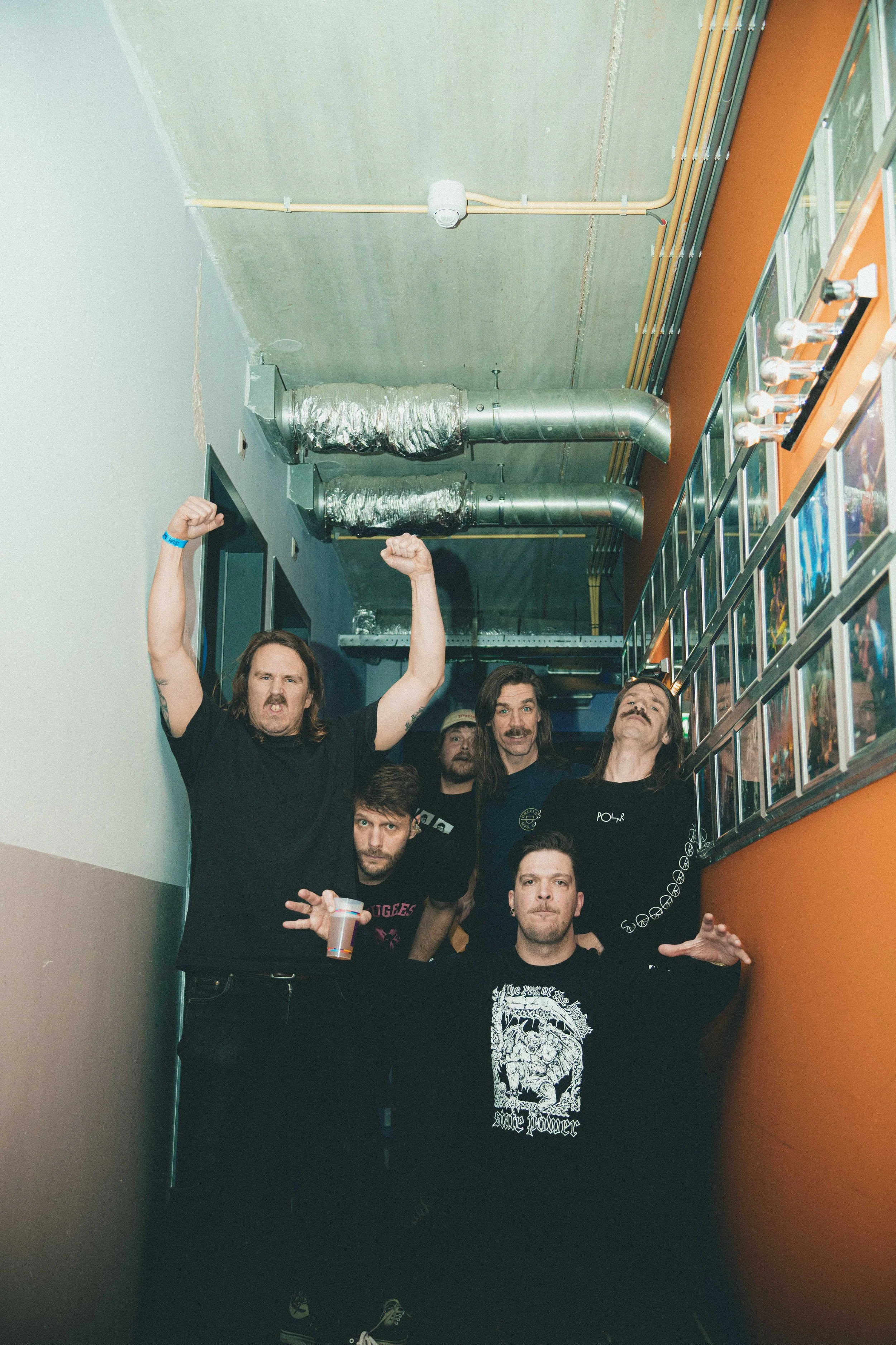Group of five men posing in a narrow hallway, some with raised fists, and one holding a drink, with framed photos on the wall and exposed ductwork on the ceiling.