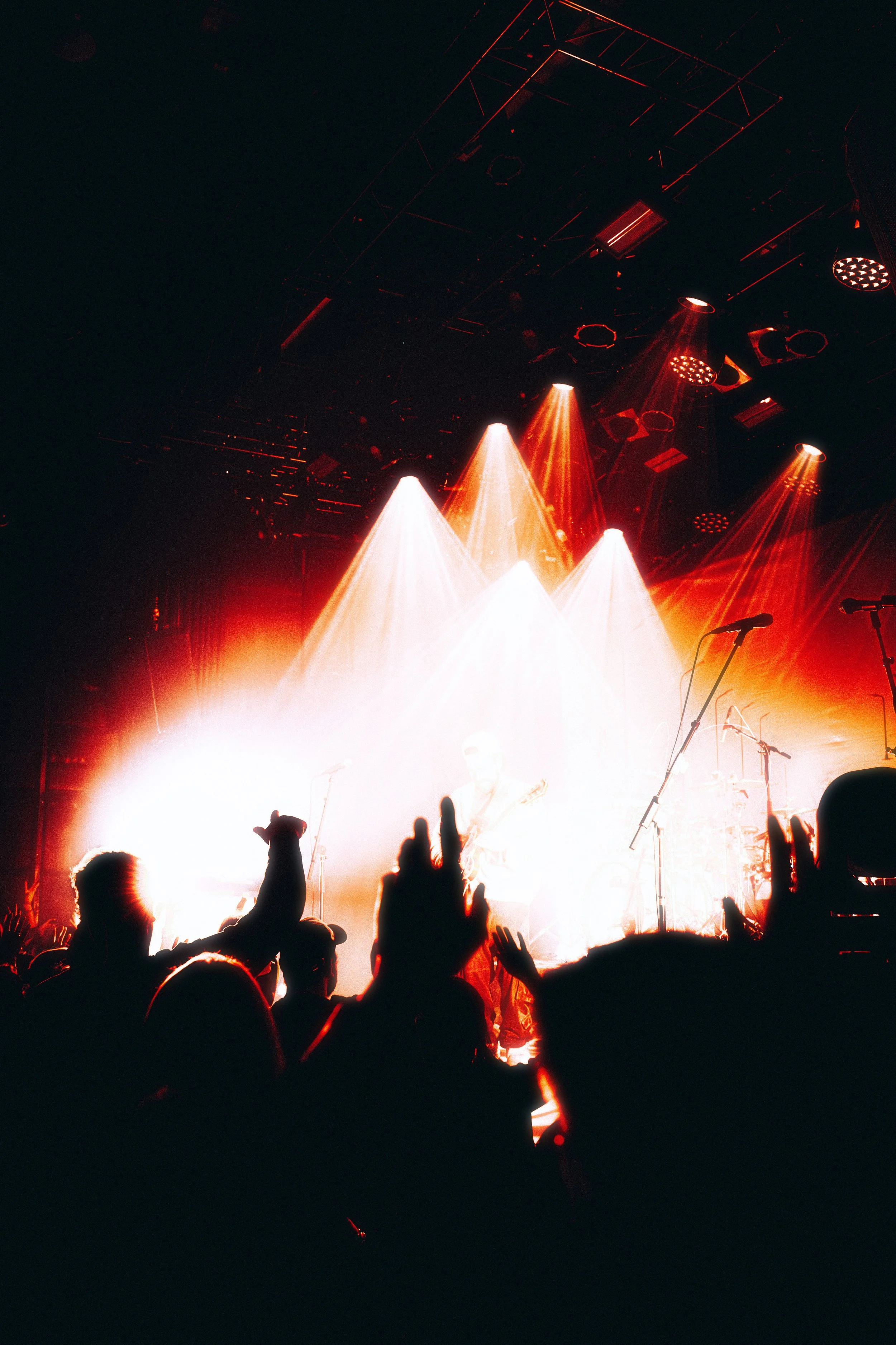 A concert stage illuminated with bright red, orange, and white lights, with a band performing and an audience in the foreground raising their hands.