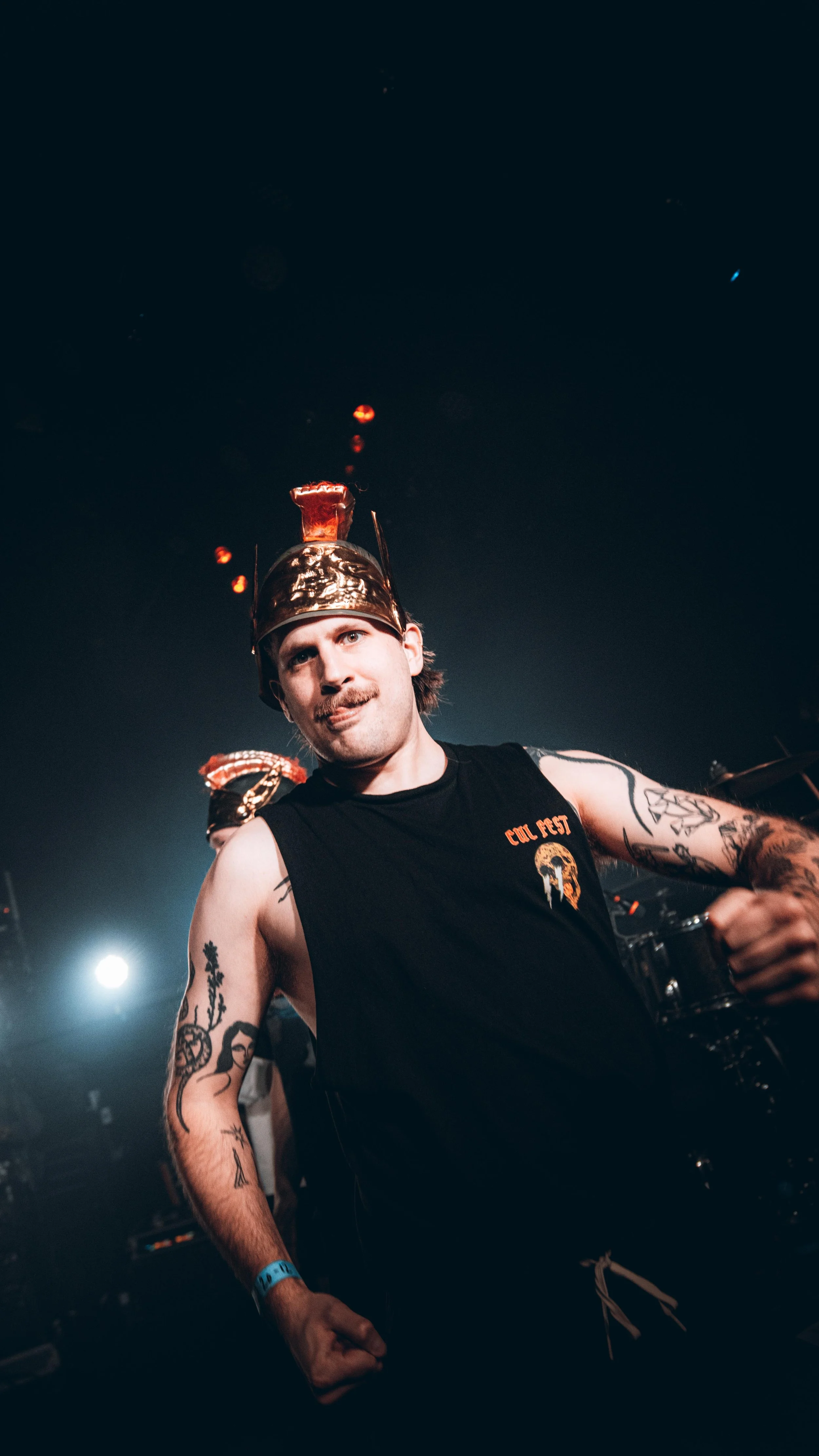 A man with tattoos on his arms wearing a black sleeveless shirt and a metallic crown with a red object on top, standing in a dark room with stage lights.