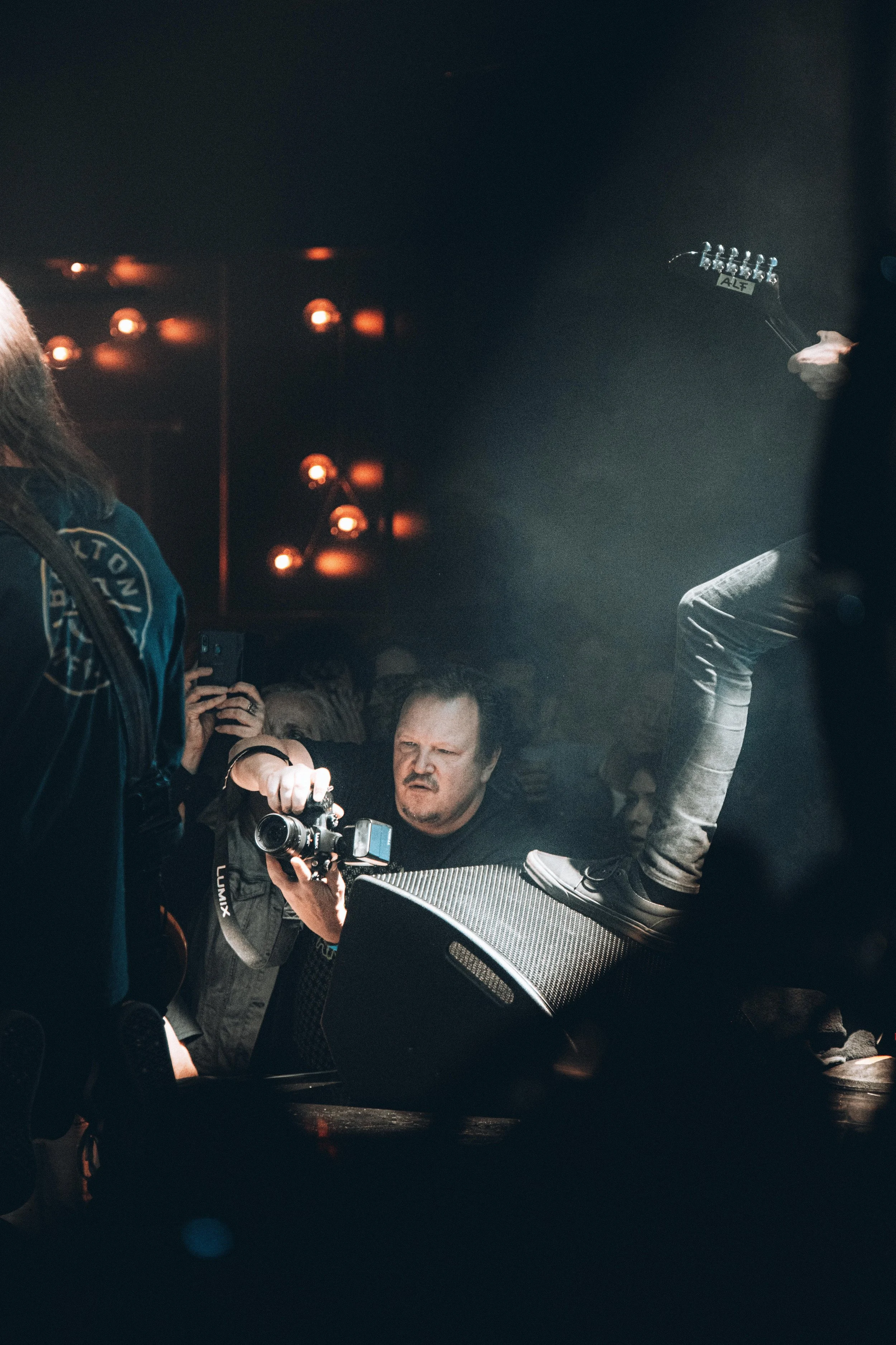 Person with long blonde hair wearing a dark jacket interacting with a photographer at a concert or event, with a crowd and stage lighting visible in the background.