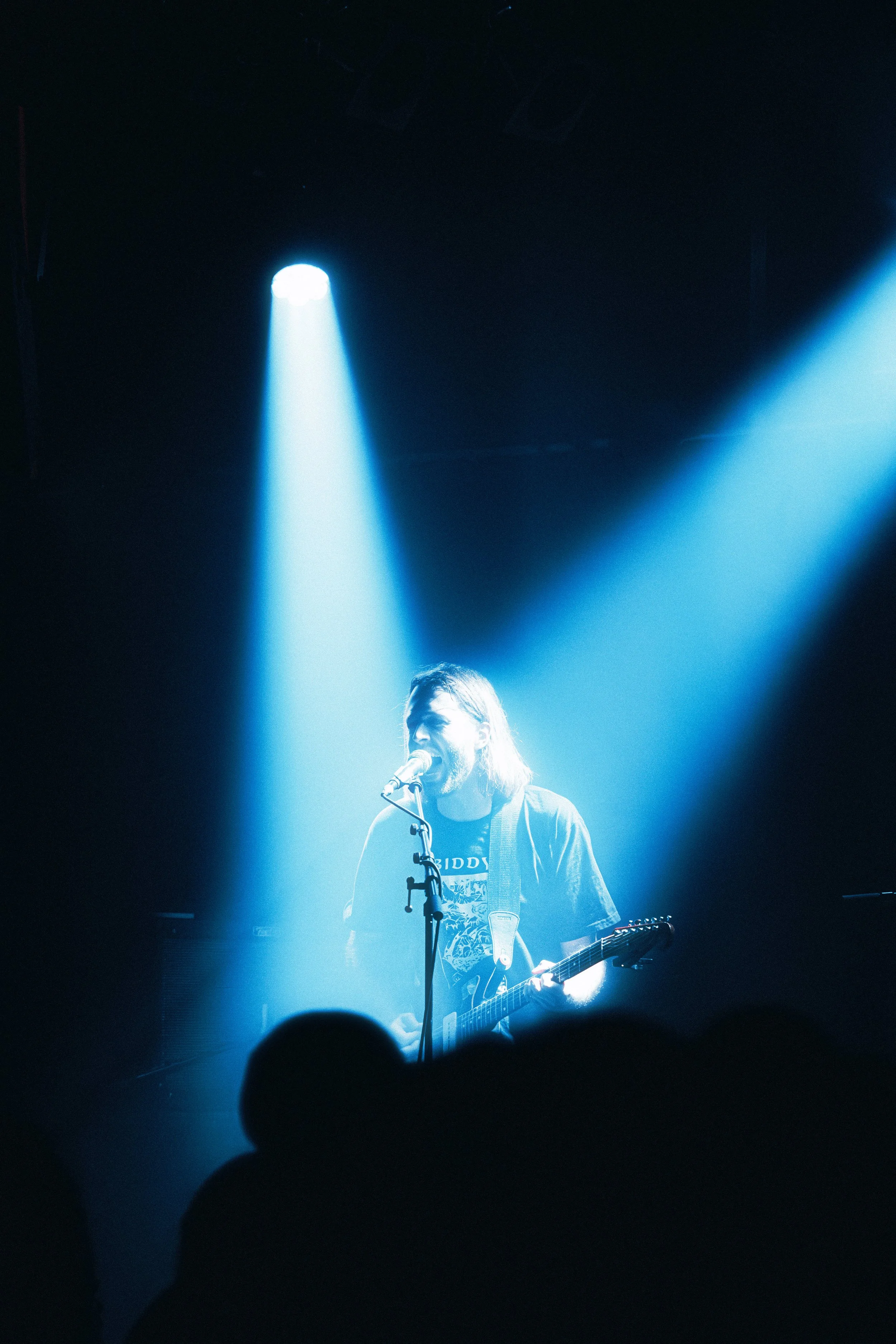 A male musician performing on stage under blue spotlight, singing into a microphone and playing an electric guitar during a concert.