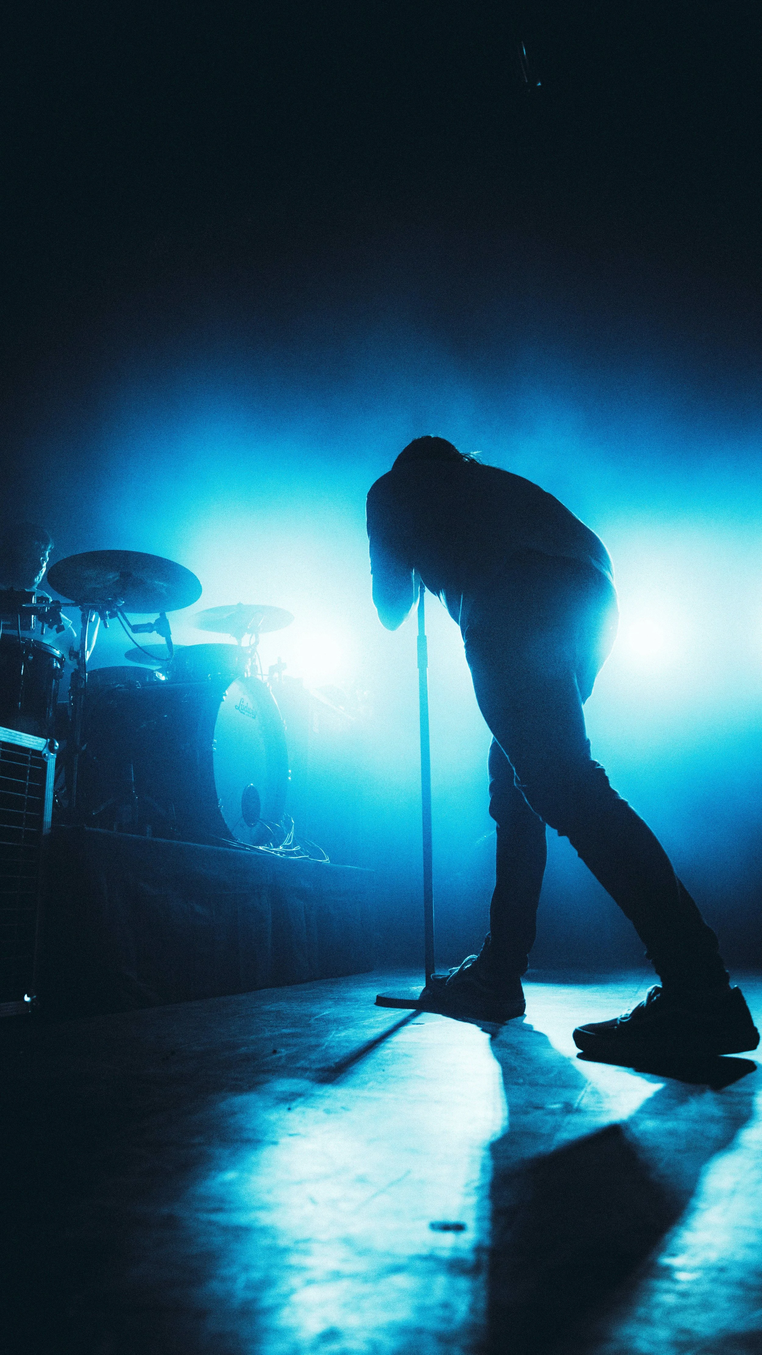 Silhouette of a person standing on a stage with a microphone, backlit by blue lights, with drums visible on the left side.