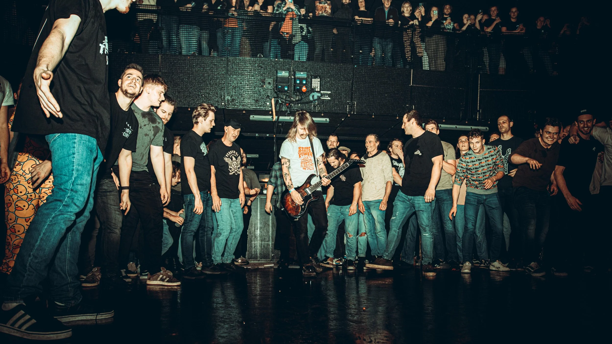 A crowd in a concert venue watching a musician with long hair and tattoos playing electric guitar on stage, surrounded by others standing and enjoying the show.