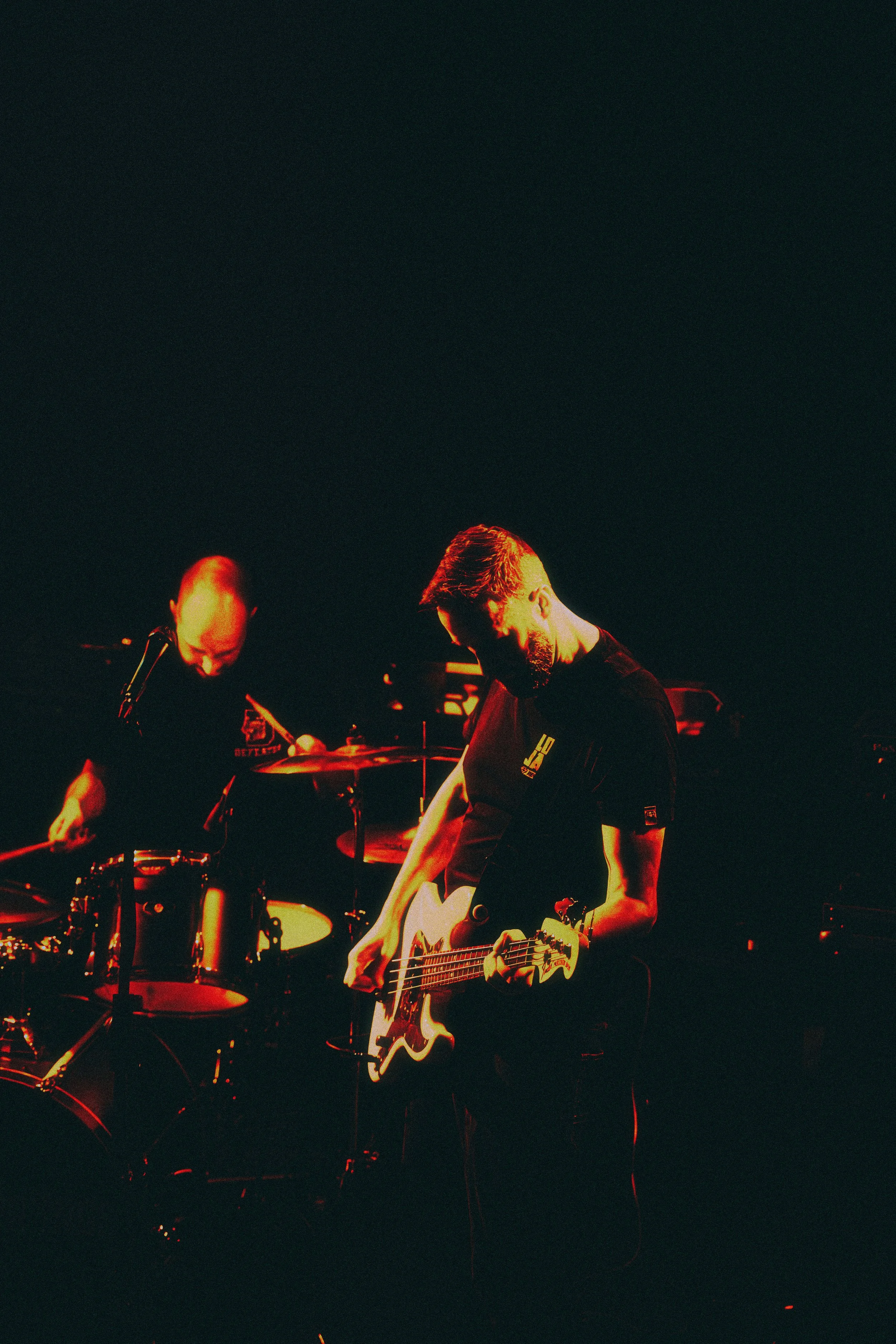 Musicians performing on stage with a guitarist and a drummer, illuminated by red stage lighting.