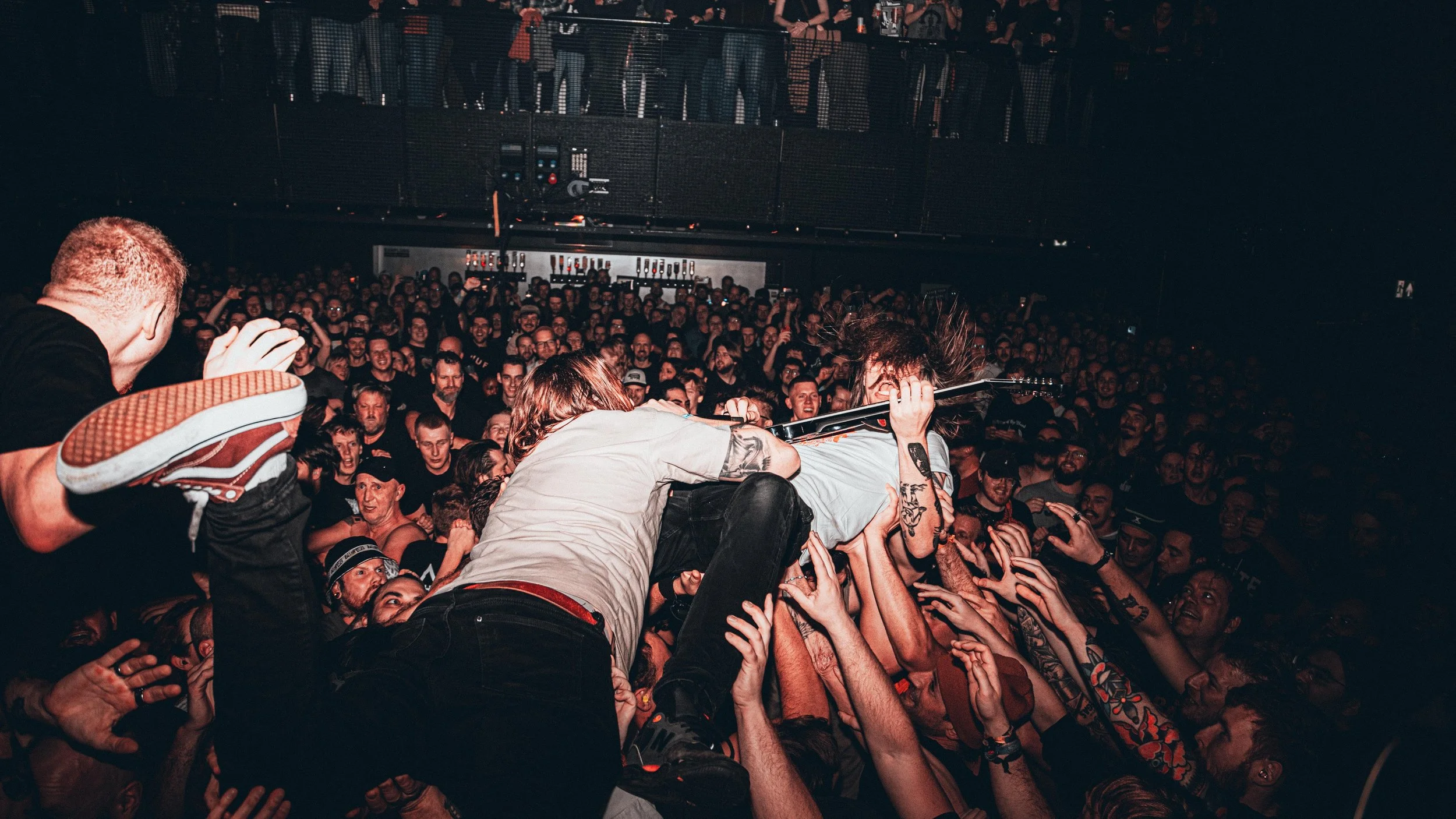 Musicians perform on stage at a concert, crowd surfing with an audience reaching out their hands, some with tattoos, in a dark concert venue.