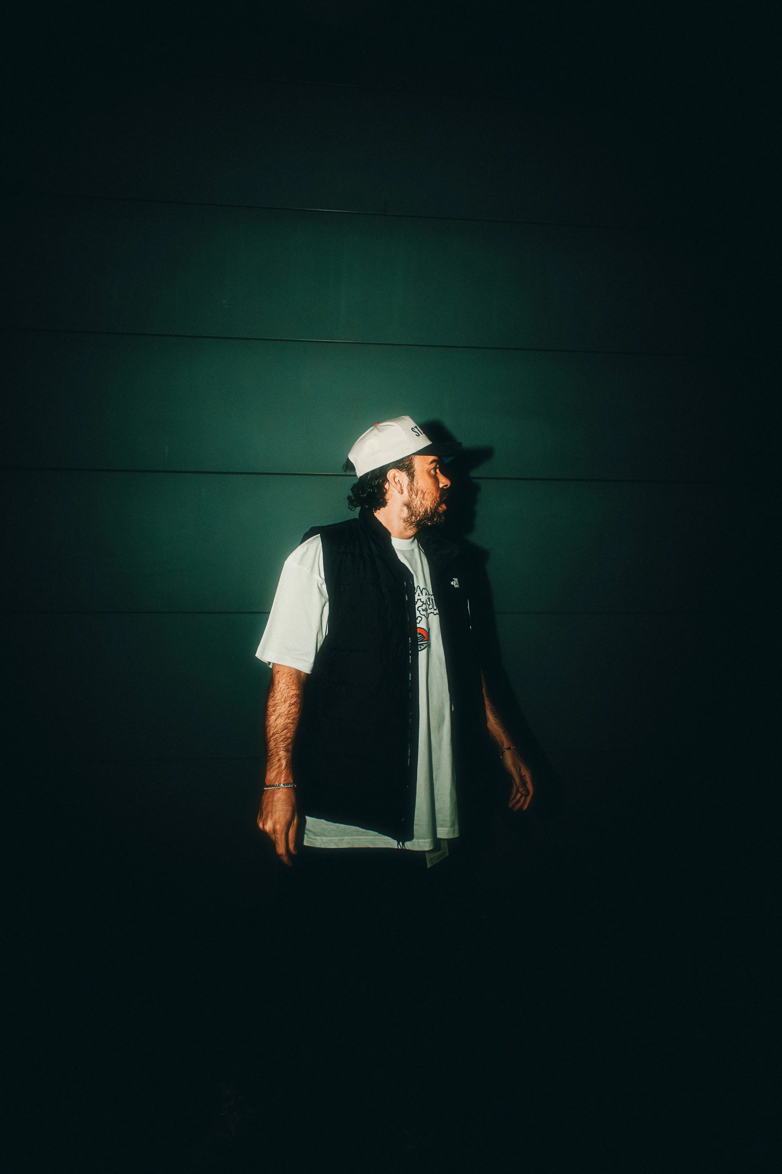A man with curly hair and a beard is standing against a dark green wall, wearing a white cap, a white t-shirt, and a black vest.