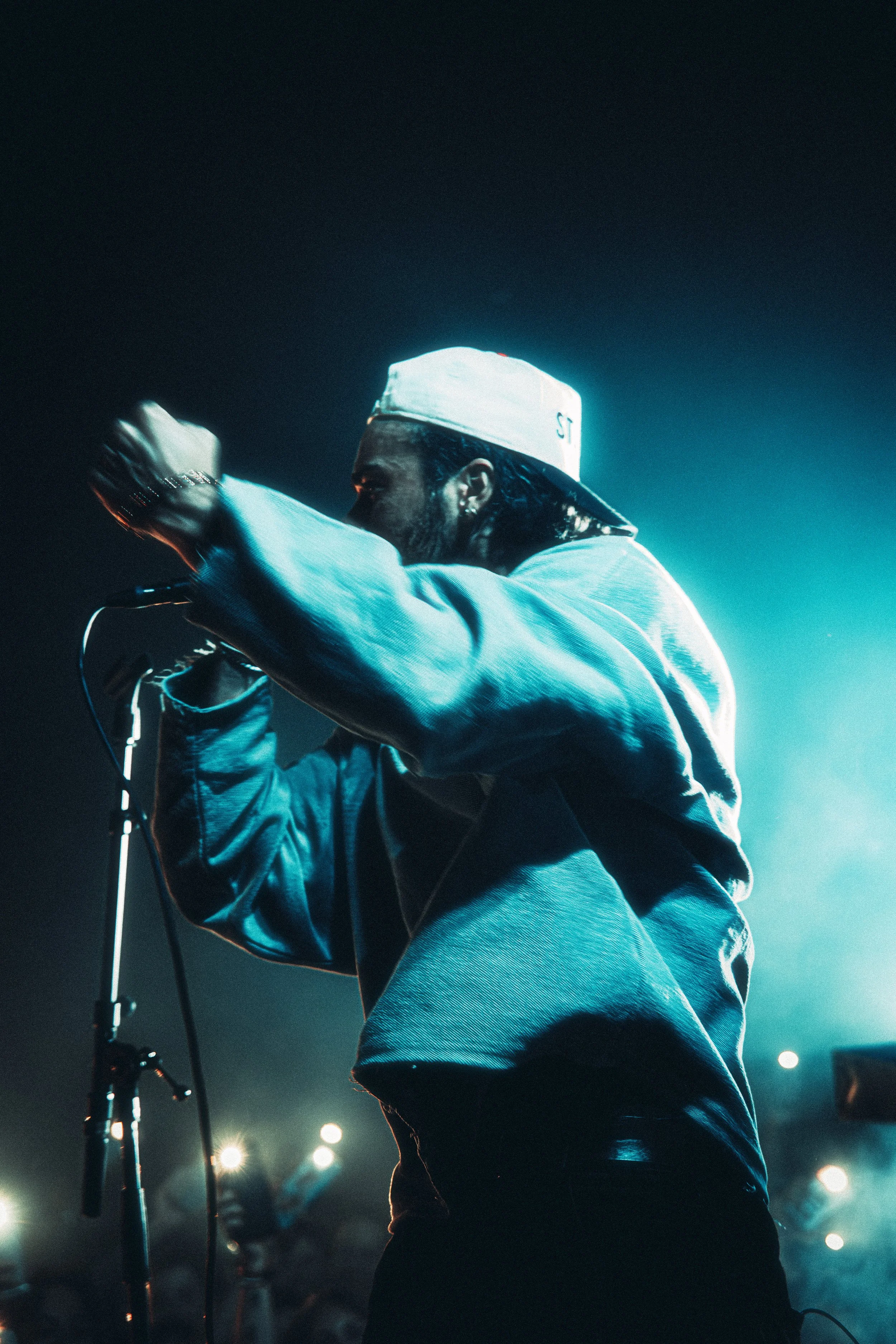 A male musician wearing a white baseball cap and a blue jacket performing on stage with microphone under blue lighting.
