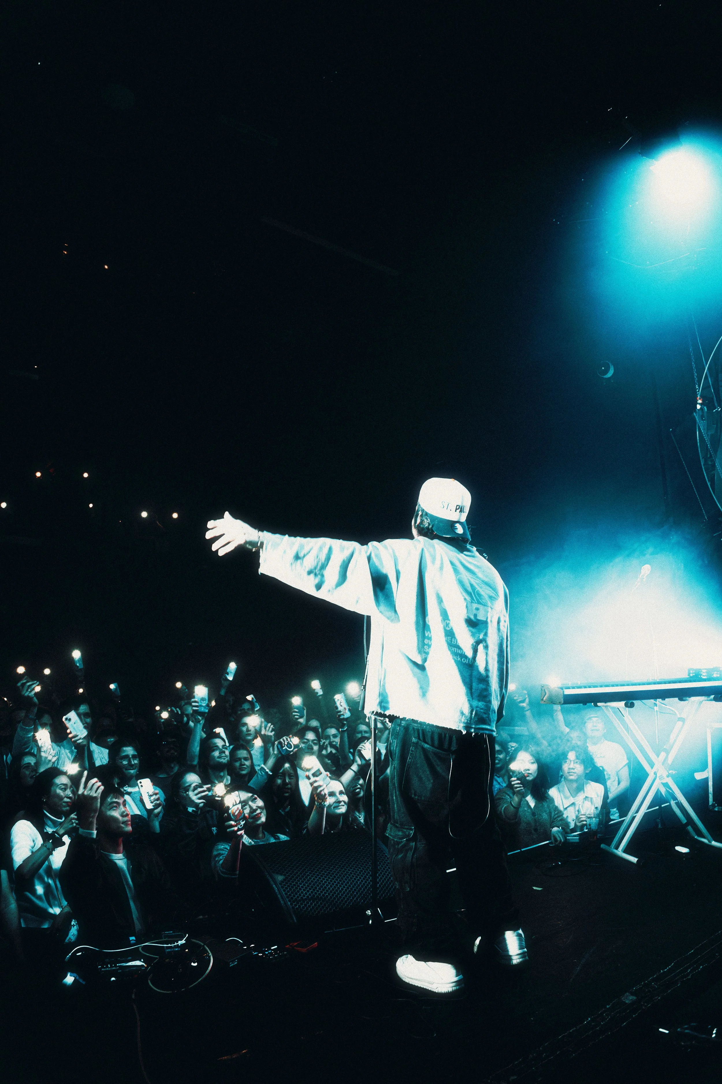 A performer with a baseball cap and casual clothing on stage facing a crowd at a concert with bright lights.