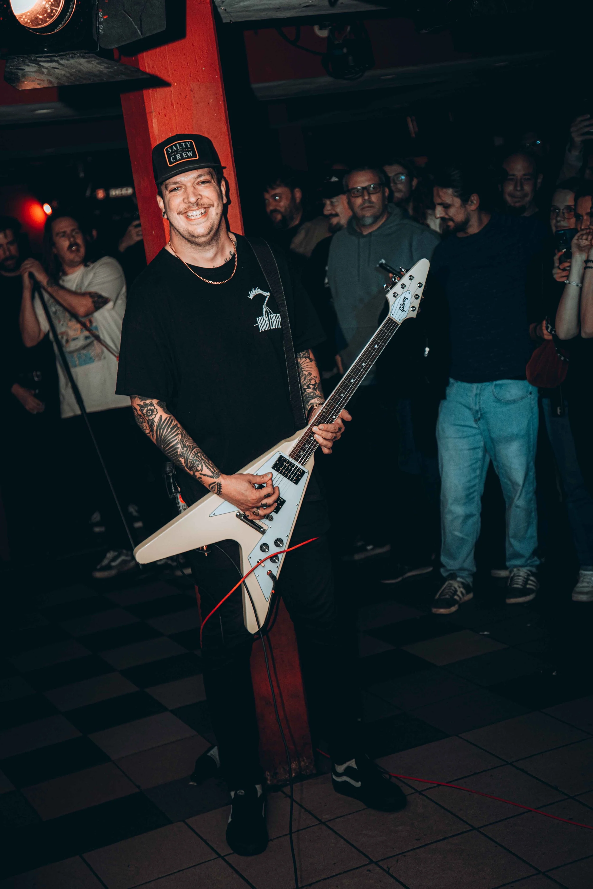 A man with tattoos on his arms and a black cap, smiling while playing a white electric guitar in a crowded indoor setting.
