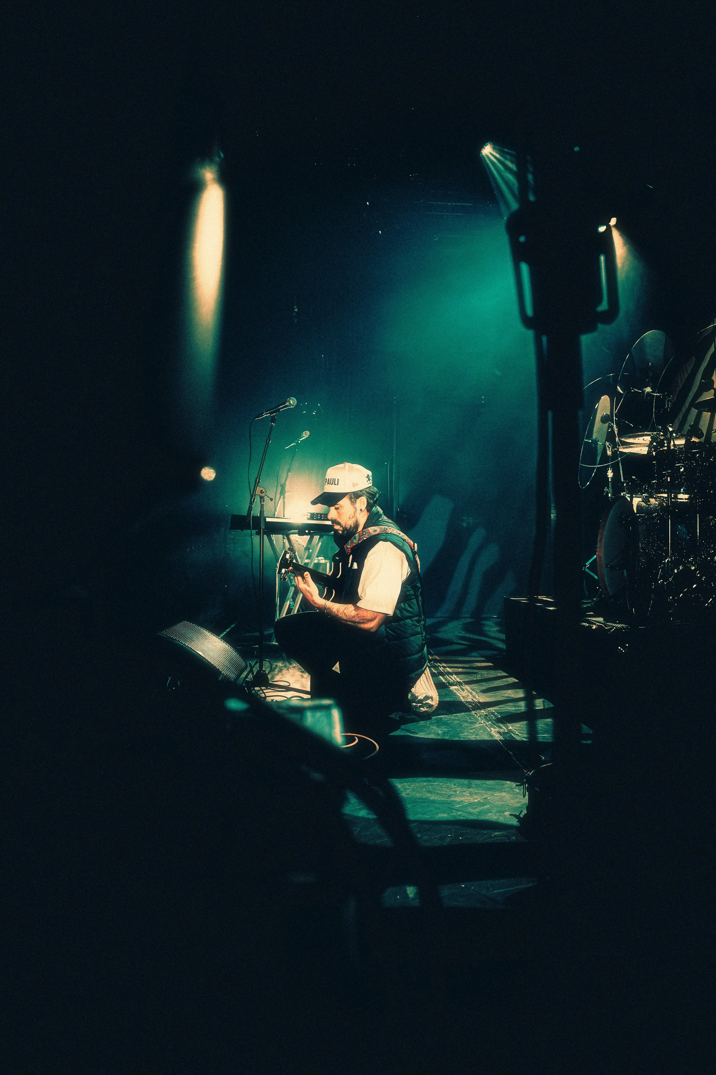 A musician kneeling on stage playing an acoustic guitar, with stage lights and musical equipment around him.