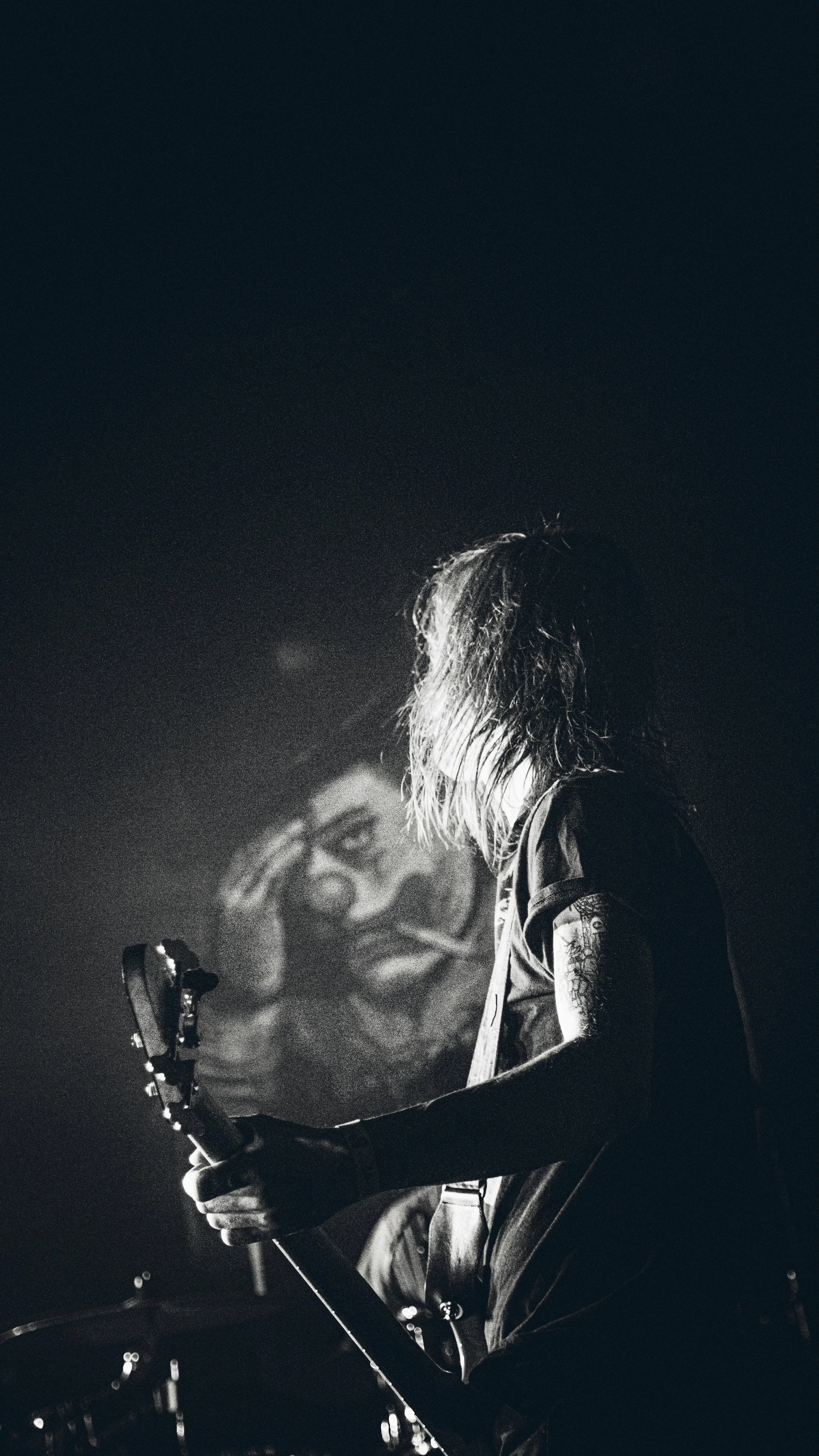 A musician with long hair playing an electric guitar on stage, with a portrait poster of a woman with sunglasses in the background.