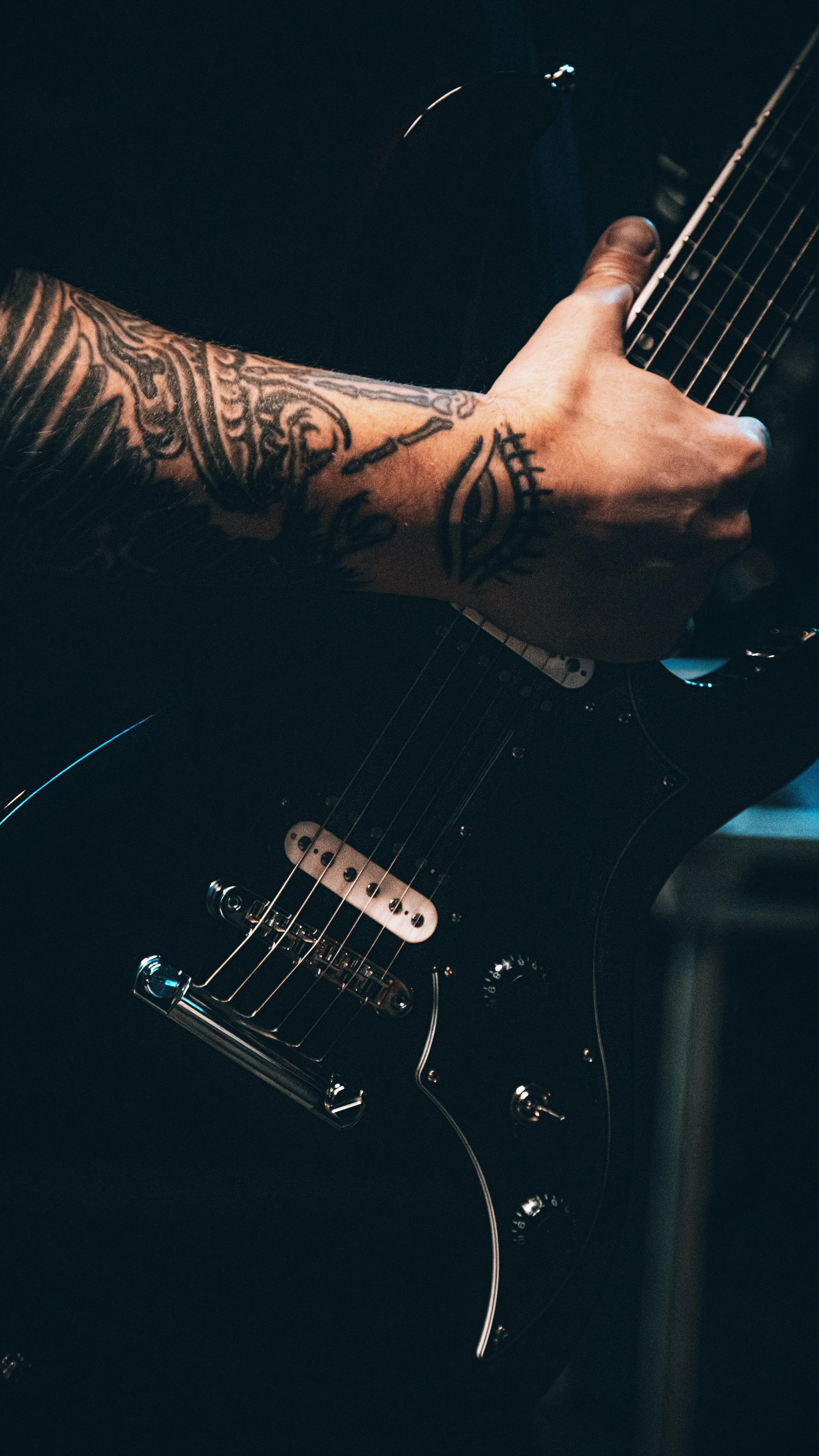 Close-up of a person with tattoos on their arm playing a black electric guitar.