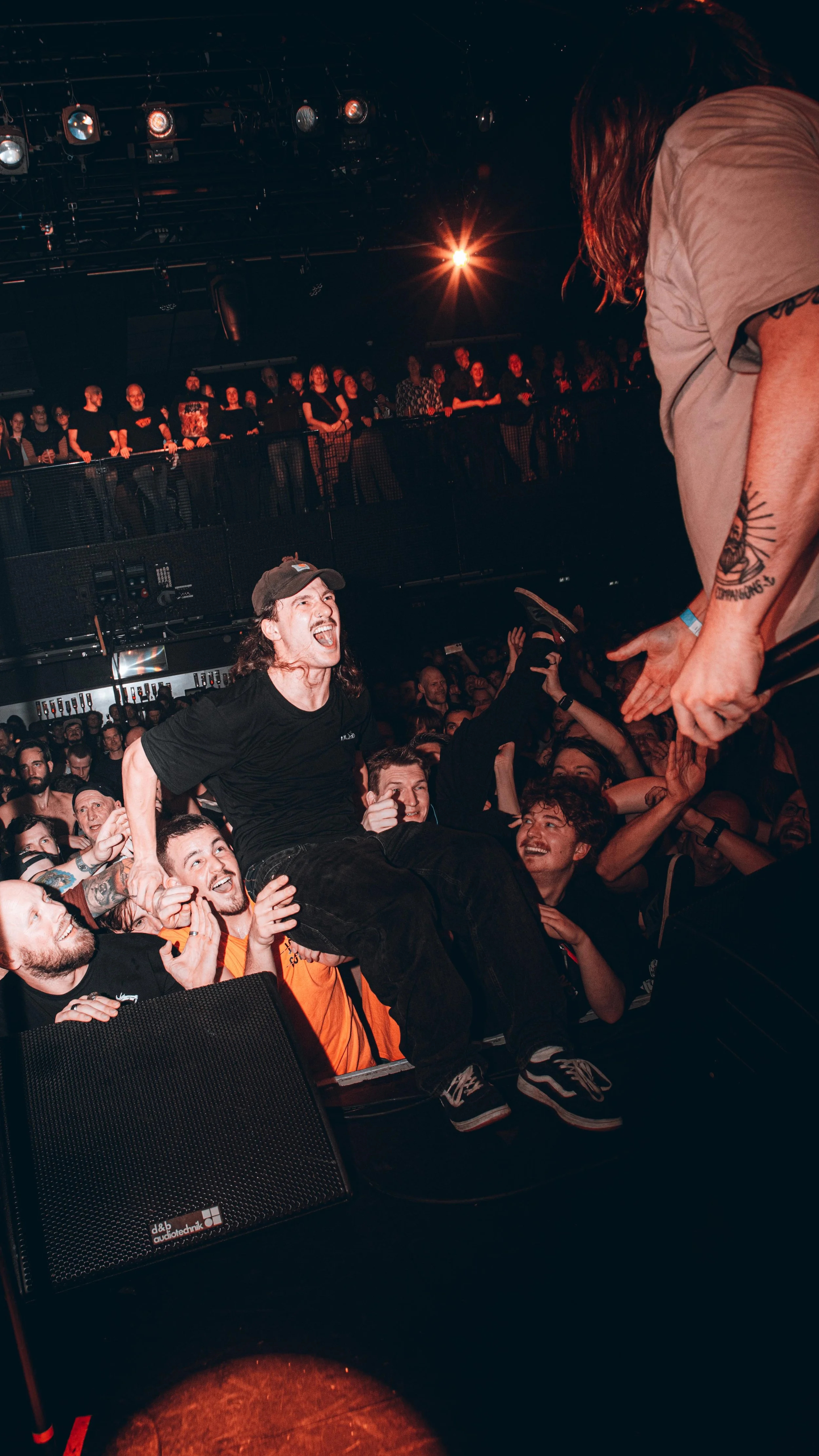 A live concert scene with a crowd enjoying the performance. A man with long hair sitting on someone's shoulders, smiling and cheering, reaching out to the performer. The audience is engaged and happy, with some people raising their hands. Stage light