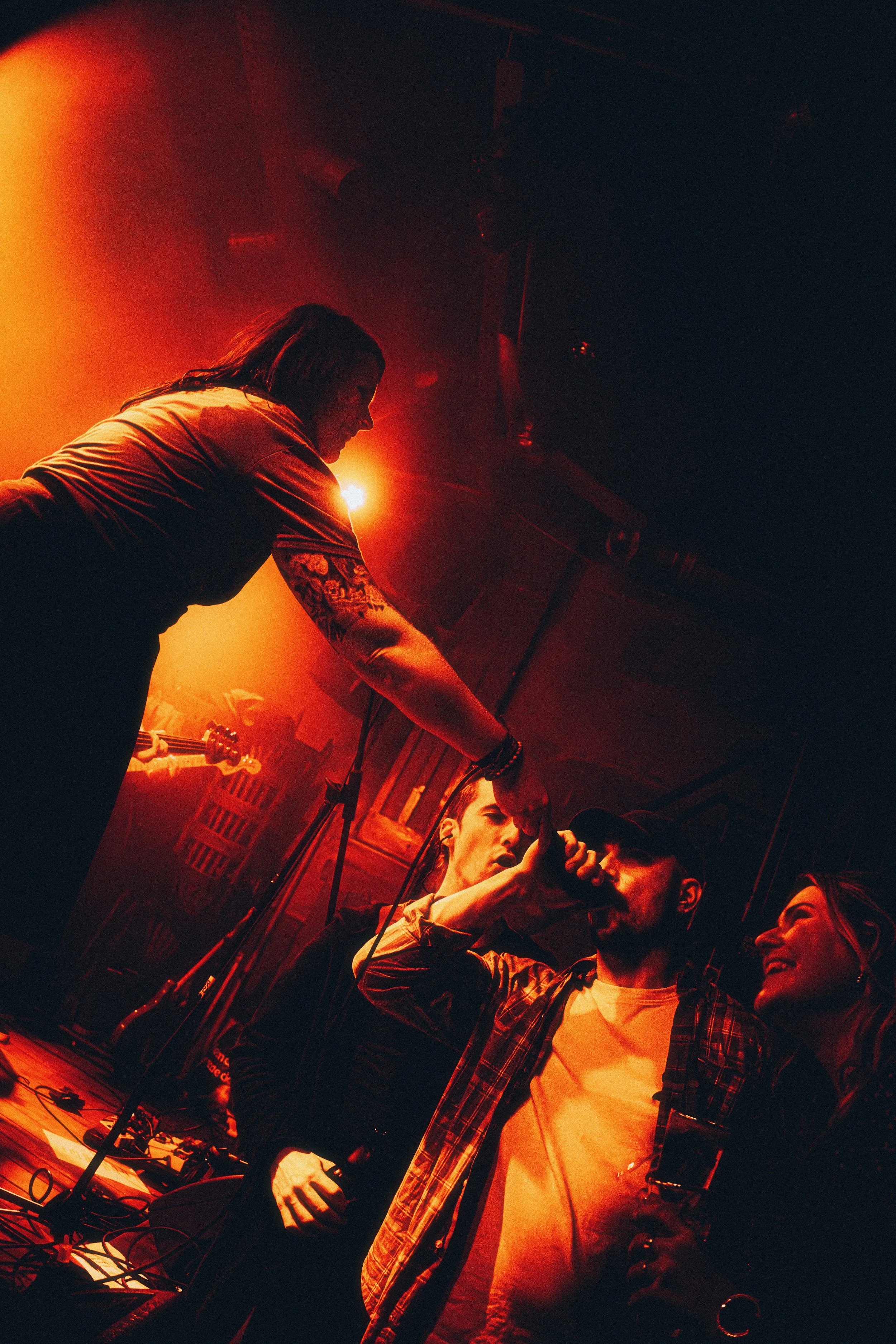 A group of people at a concert or party, with a woman on stage reaching out to a man in the crowd, who is holding a microphone, with warm orange lighting.