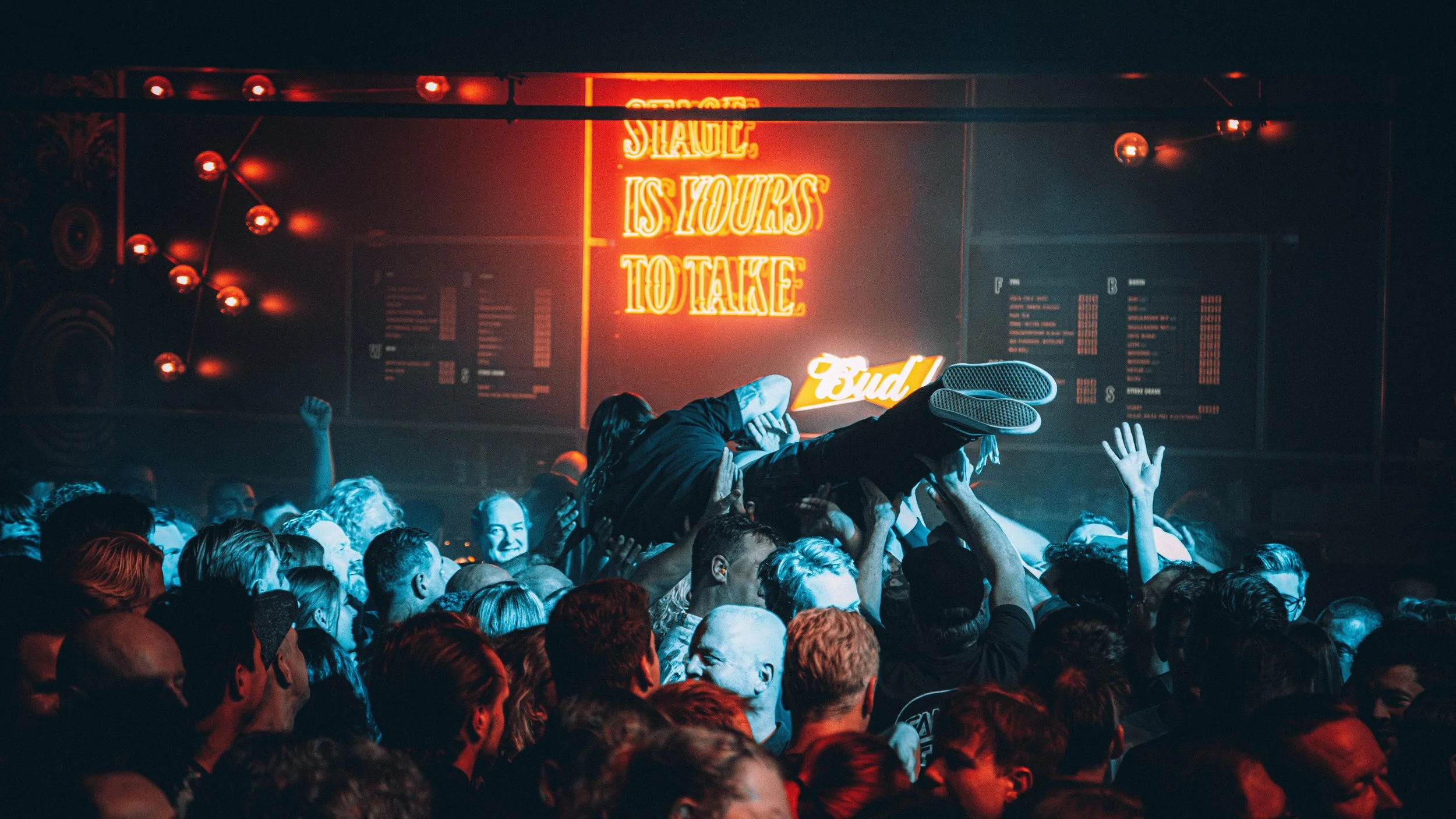 A crowd at a concert or nightclub holding a person in their arms above their heads. Neon sign in the background reads 'Shake, is hours to take.'