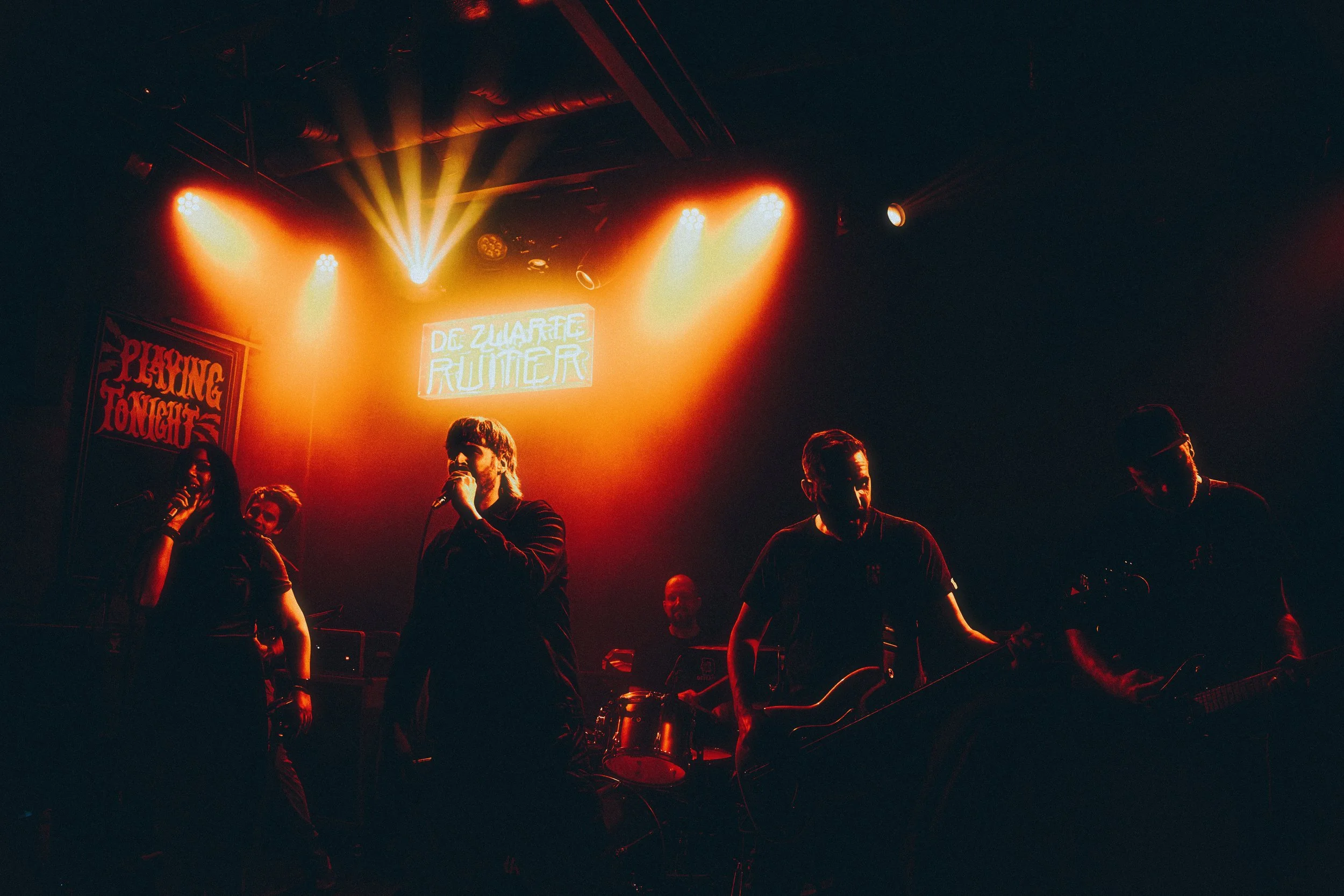 A band performing on stage with orange stage lights and a sign that says 'Playing Tonight' and 'De Zwarte Ruiter' in the background.