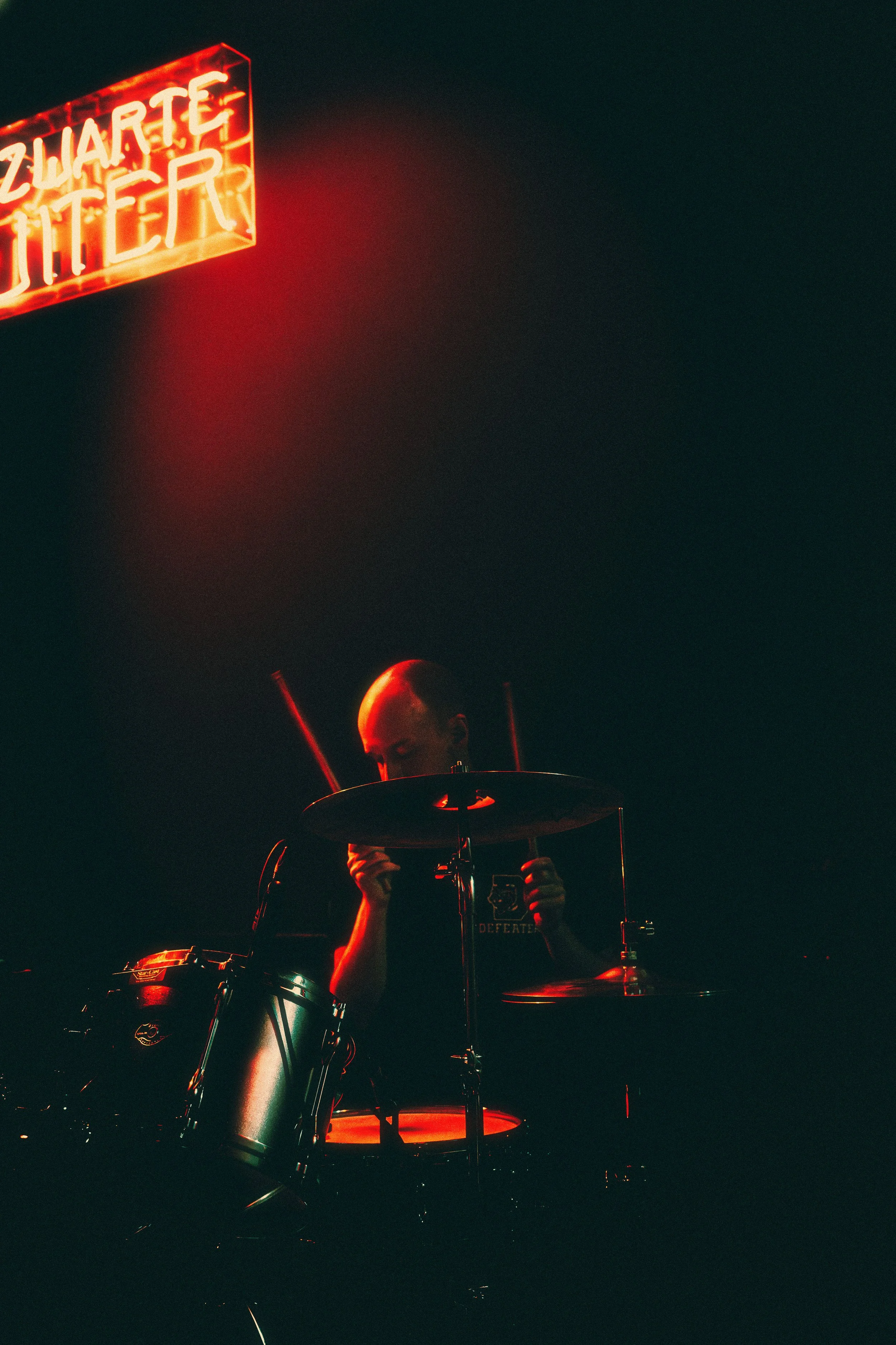 Person playing drums in dim lighting with a neon sign reading 'Zwarte Titer' above.