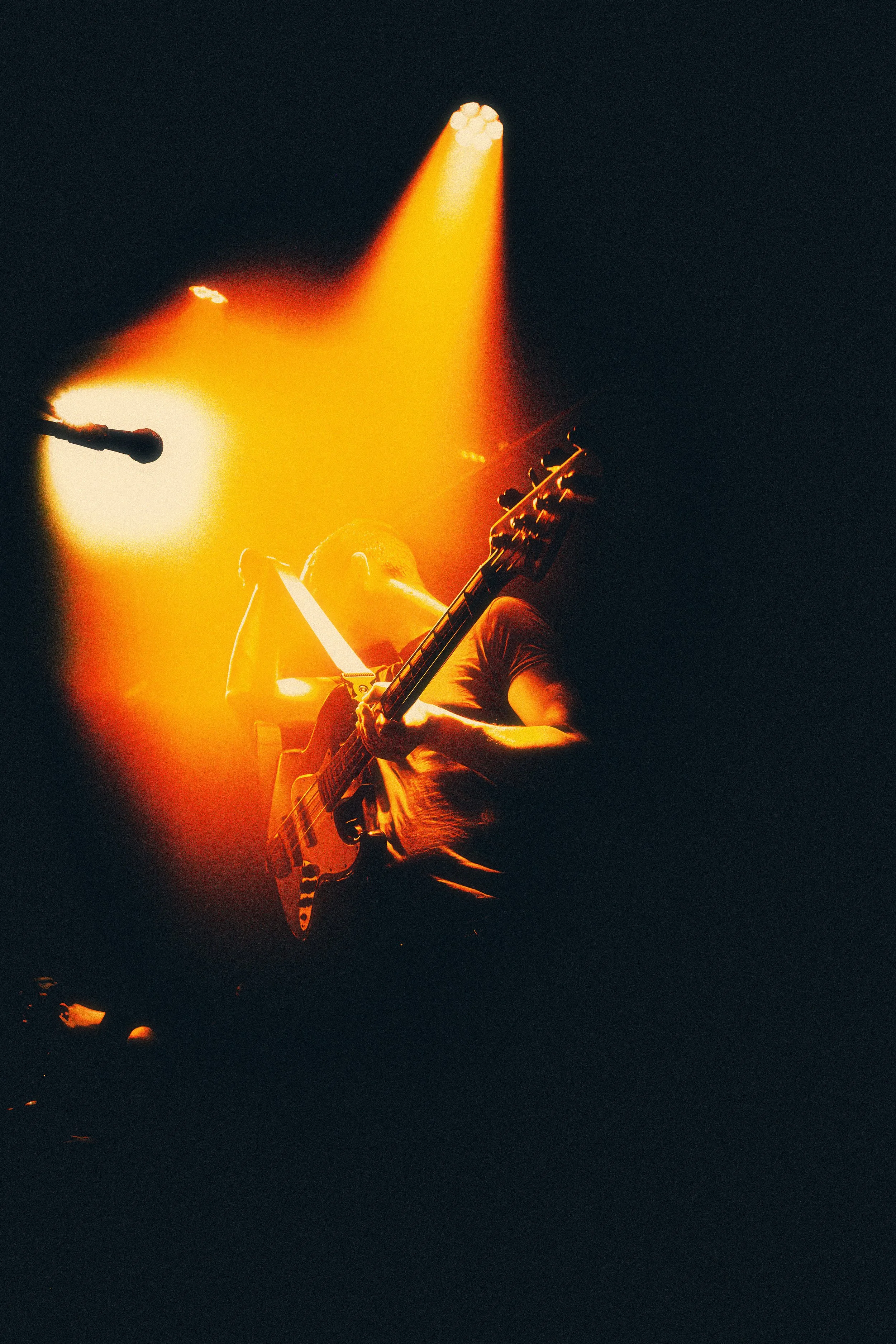 Musician playing an electric guitar on stage, illuminated by orange and yellow stage lights, with a microphone nearby in a dark setting.