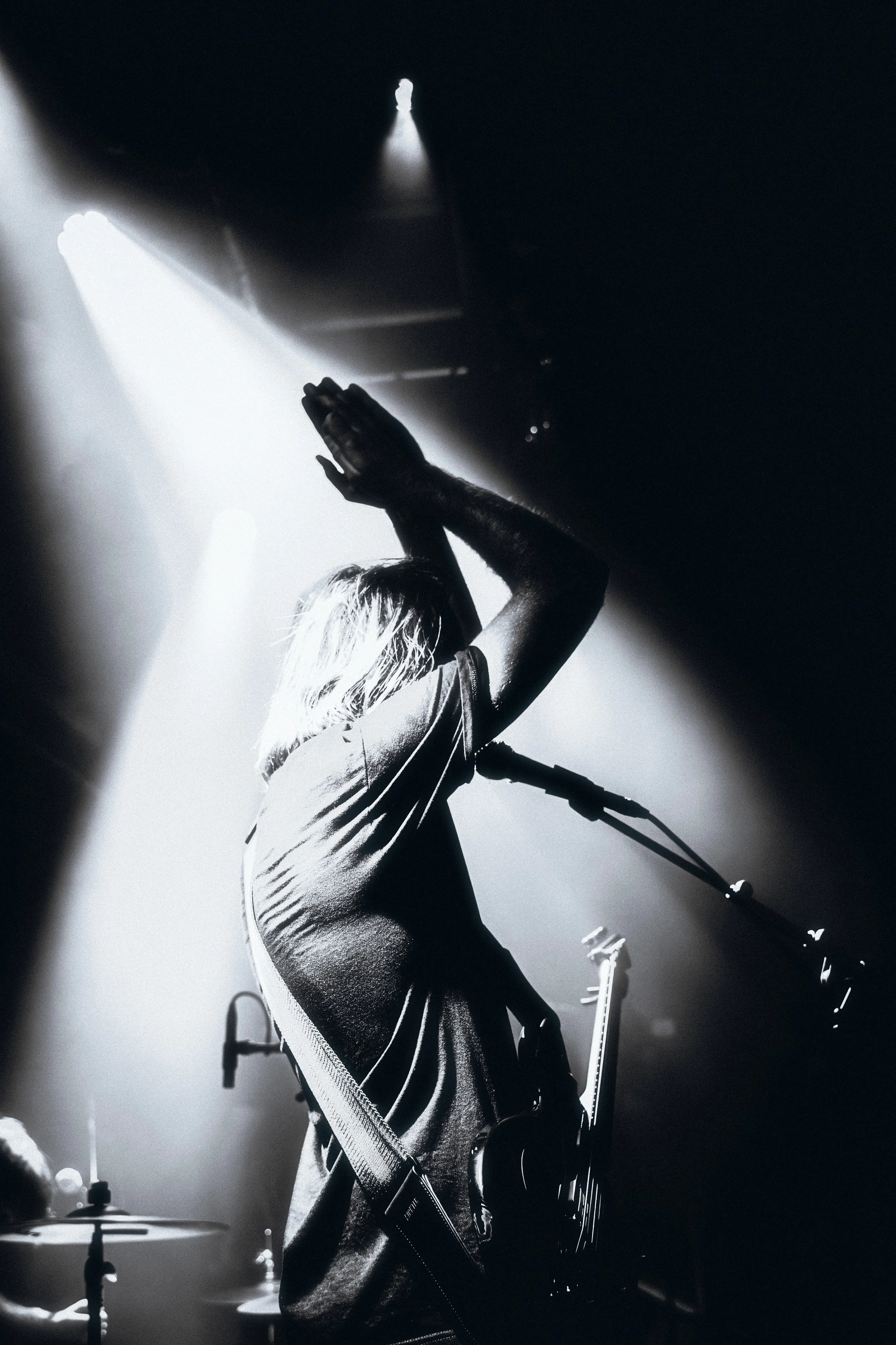 A musician on stage with a guitar, backlit by bright stage lights, raising their hand in the air during a performance.