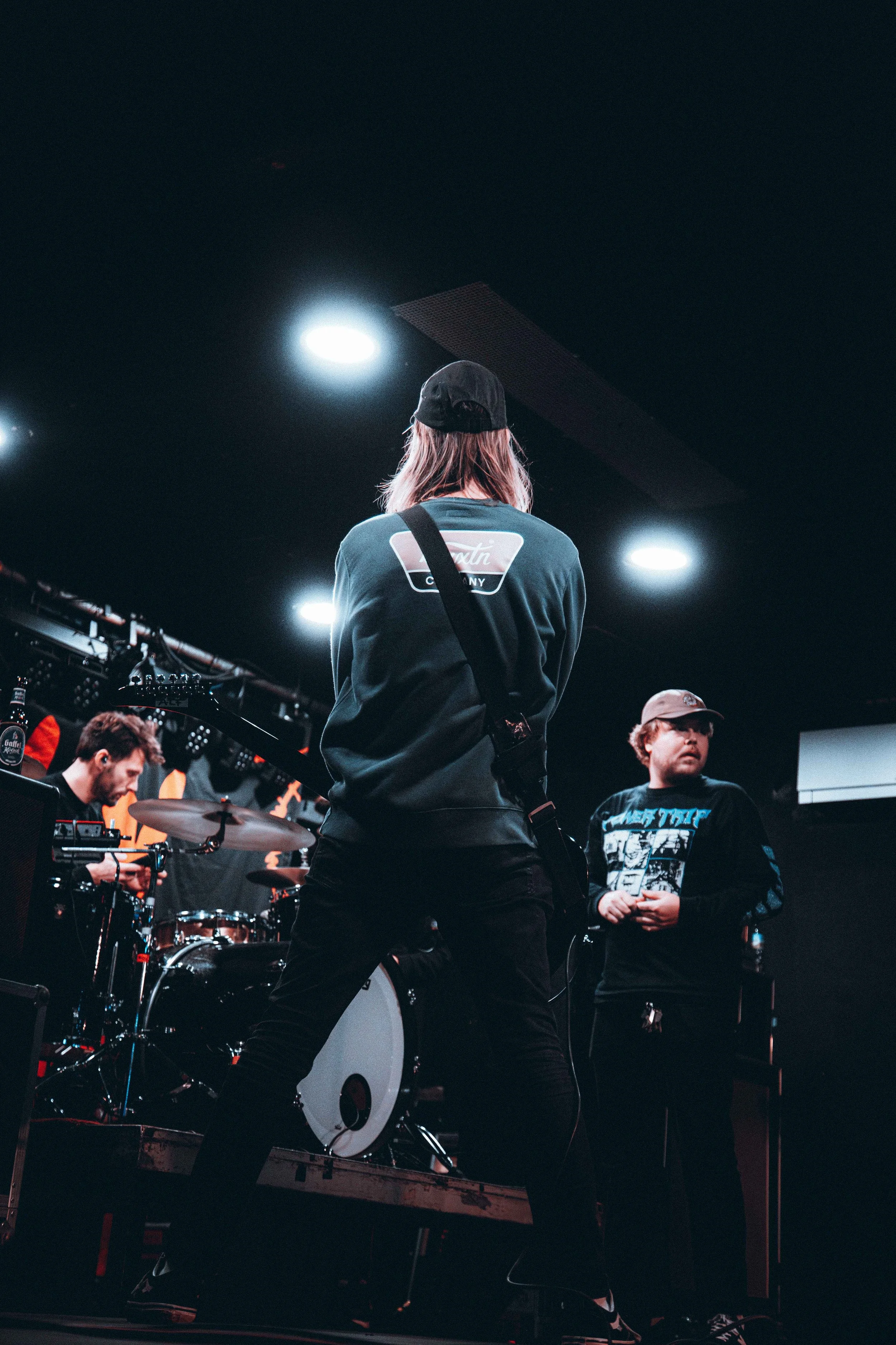 A band performing on stage with musicians playing instruments, including drums, under stage lights in a dark setting.