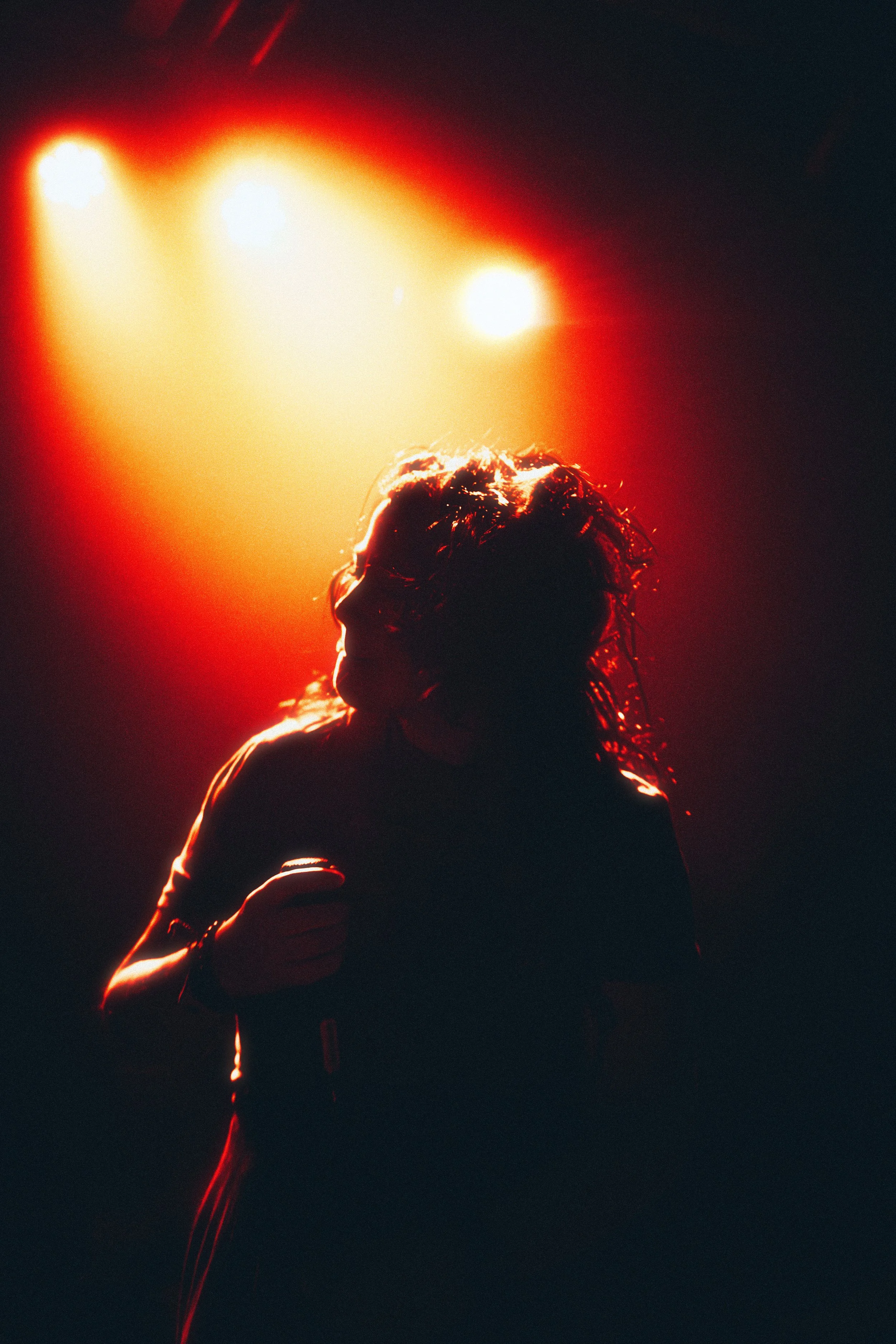 Silhouette of a person with long hair holding a microphone, backlit by red and yellow stage lights in a dark venue.