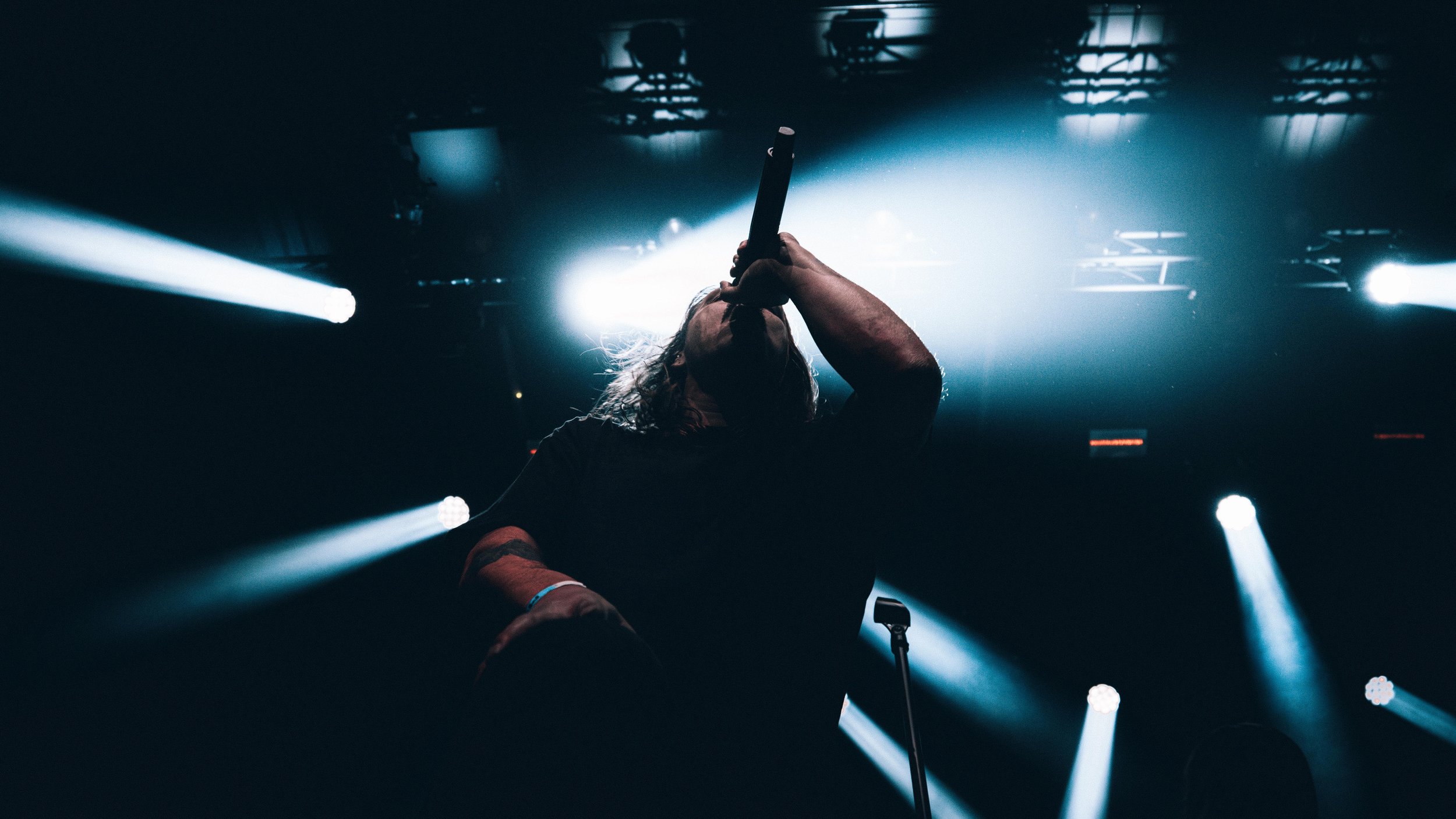 A performer with long hair on stage, holding a microphone up to their mouth, with bright stage lights shining behind them in a low-angle shot.