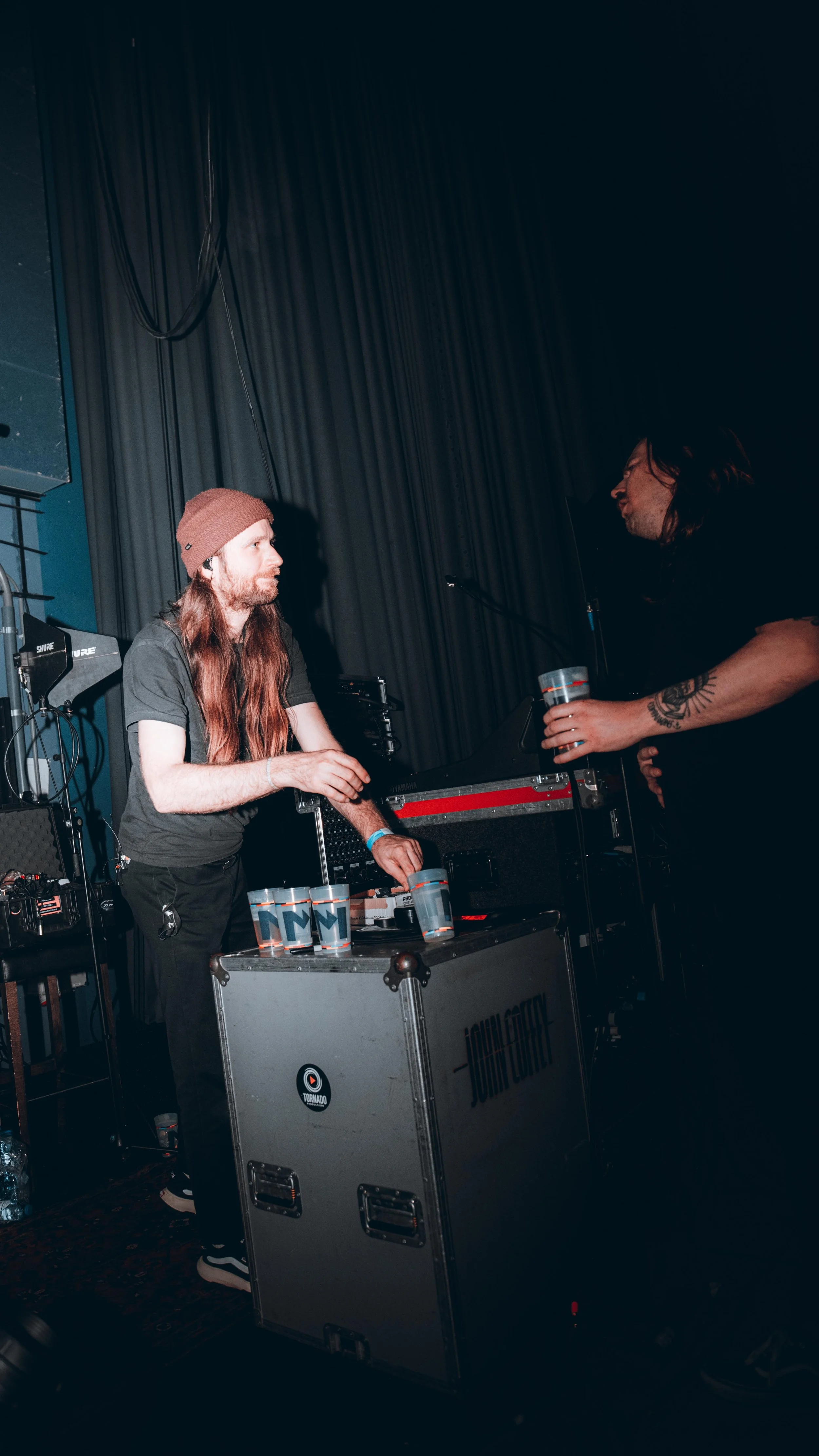 A man with long red hair, wearing a brown beanie and black t-shirt, stands behind a DJ setup with multiple CDs, while another person hands him a drink in a plastic cup. The background includes a black curtain and music equipment.