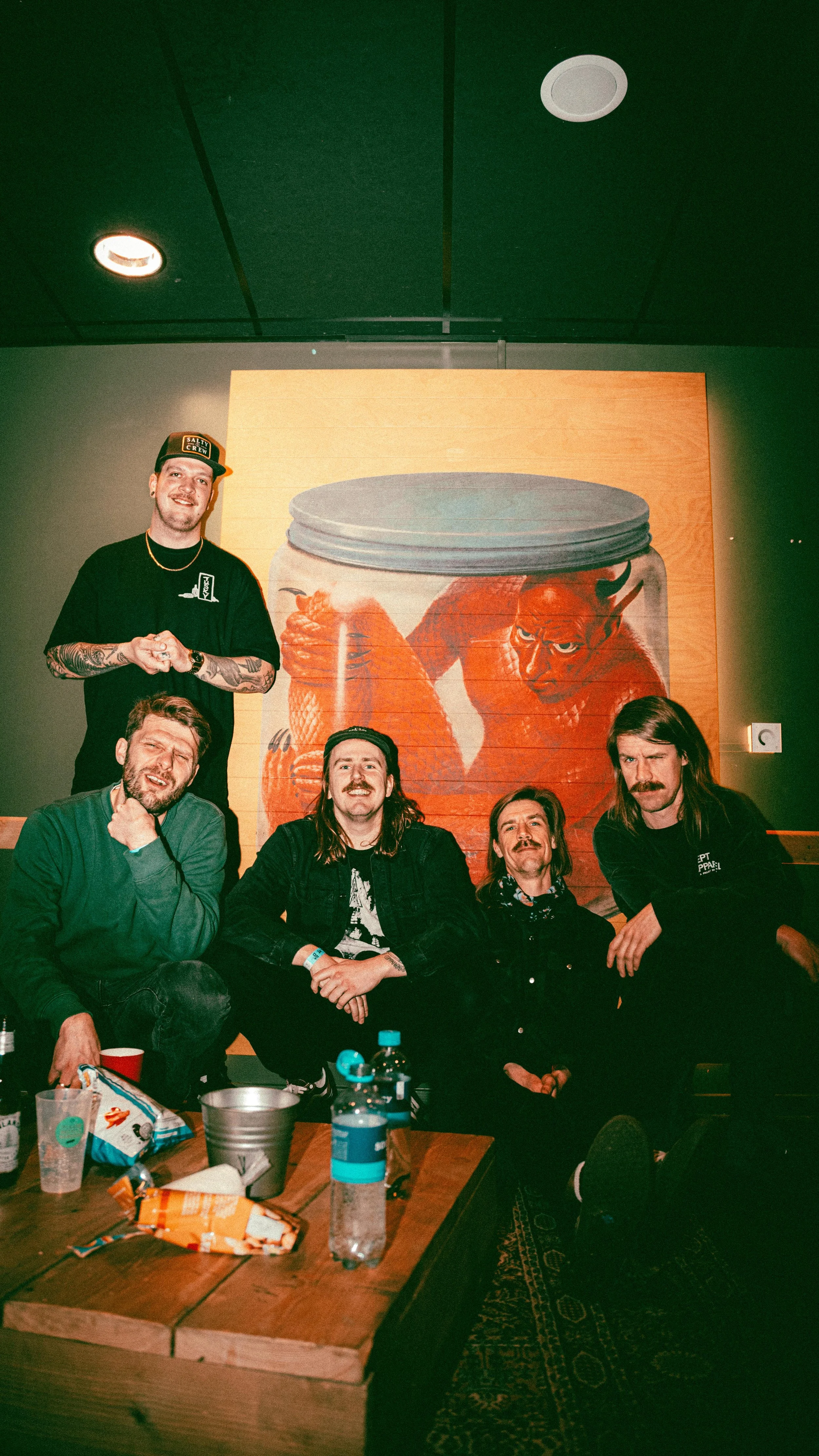 Five men with various hairstyles and facial hair, posing in front of a large wall mural of a red devil and fish inside a glass jar. Four men are sitting and one man is standing, all dressed casually in dark clothing. There are snacks and drinks on th