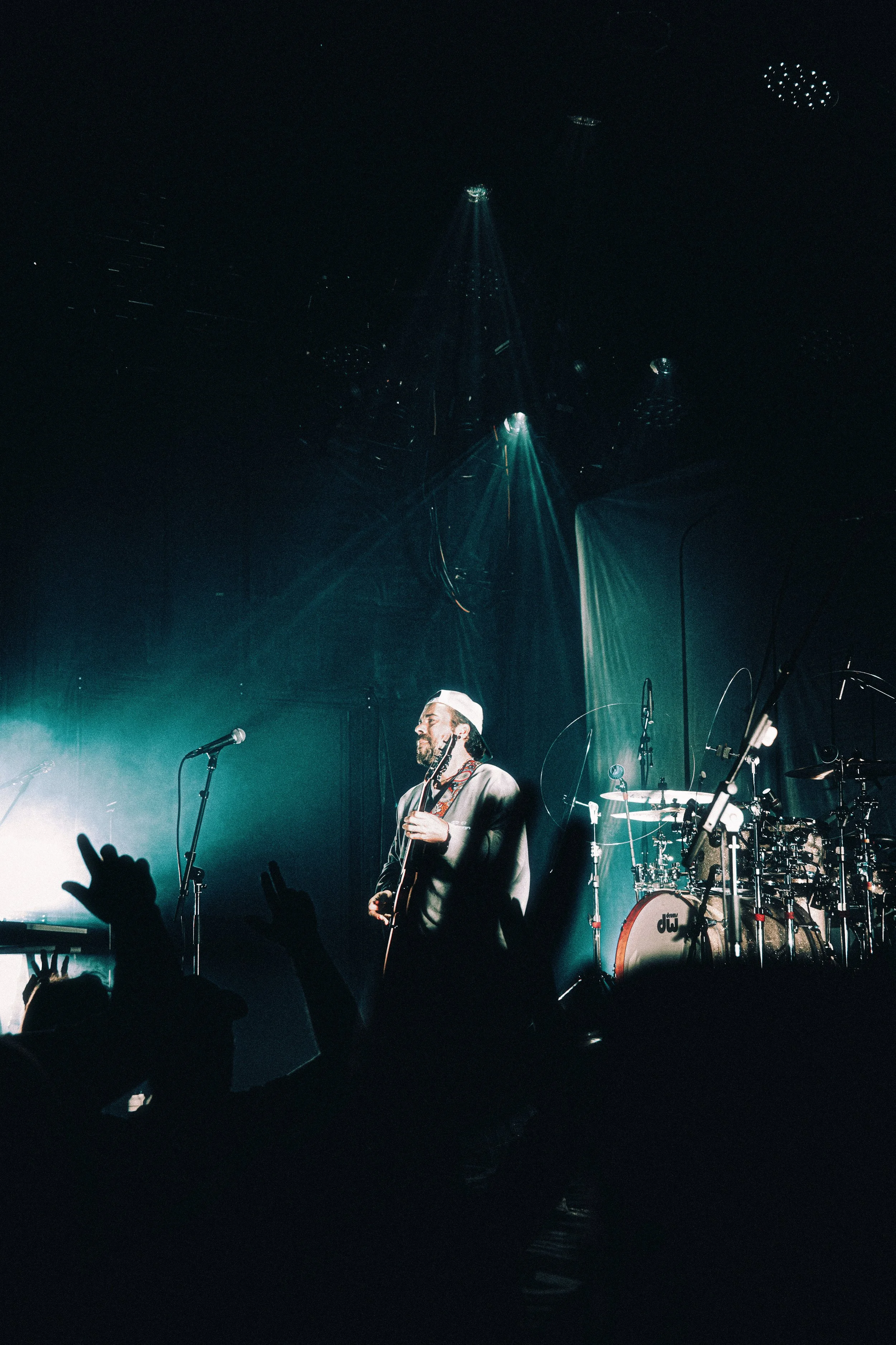 A musician performing on stage with a guitar, microphone, and drum set illuminated by stage lights. Audience members' hands are raised in the foreground.