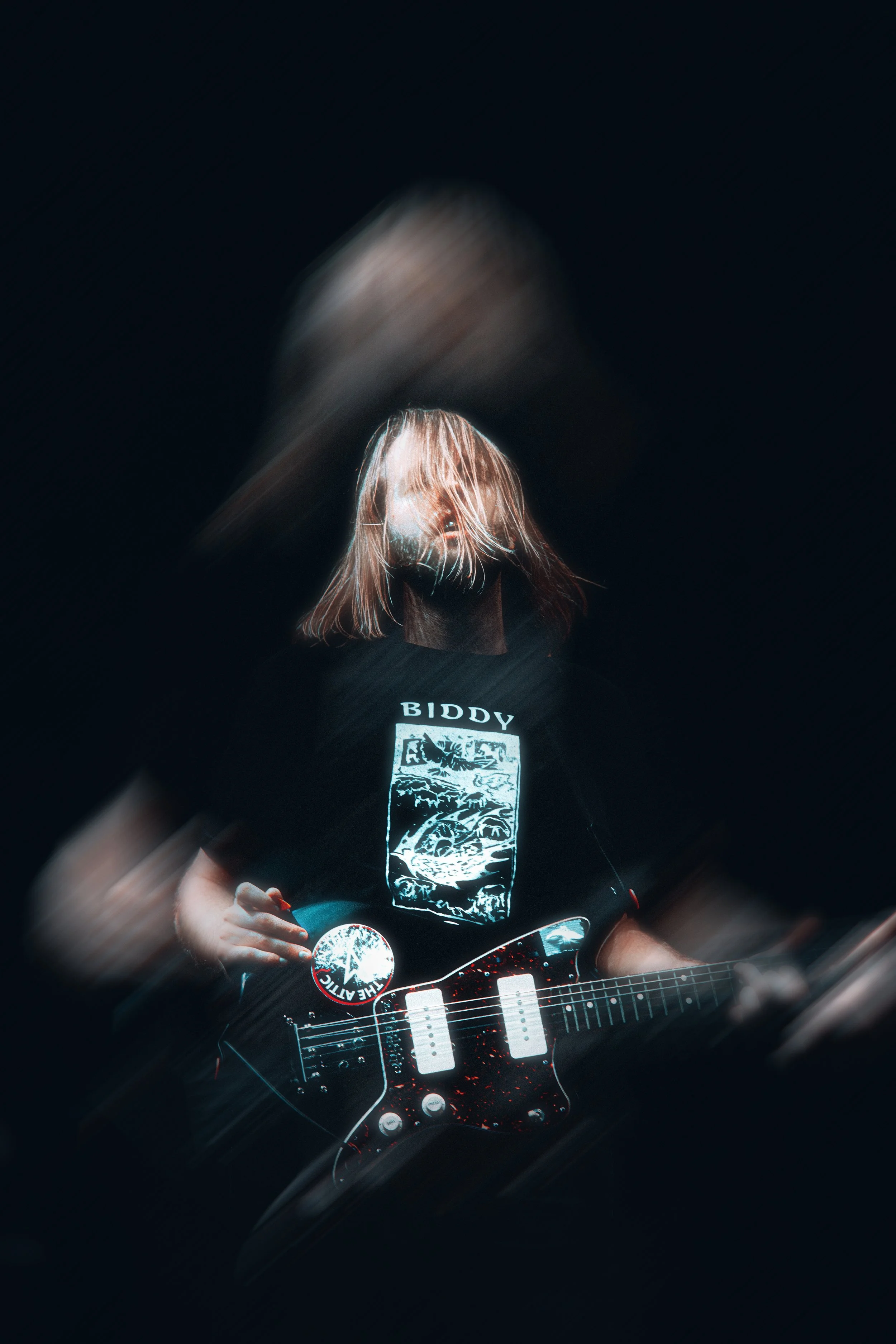 A person with shoulder-length blonde hair wearing a black T-shirt with a graphic design and white text, holding a black electric guitar with red splatter pattern, against a dark background, with motion blur and light streaks.