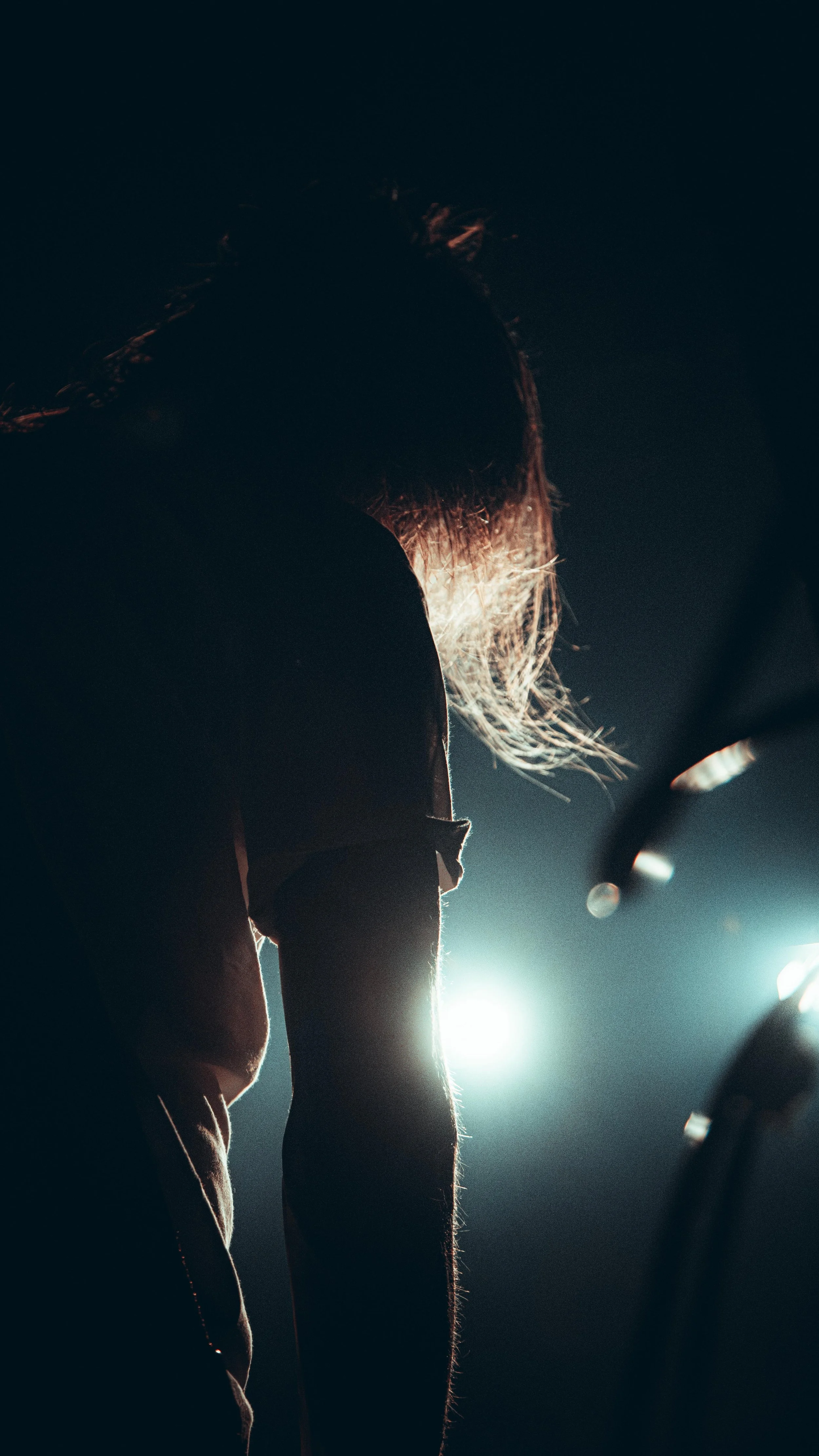 A person's silhouette bending over in a dark environment with bright light behind them, highlighting their hair and arm.