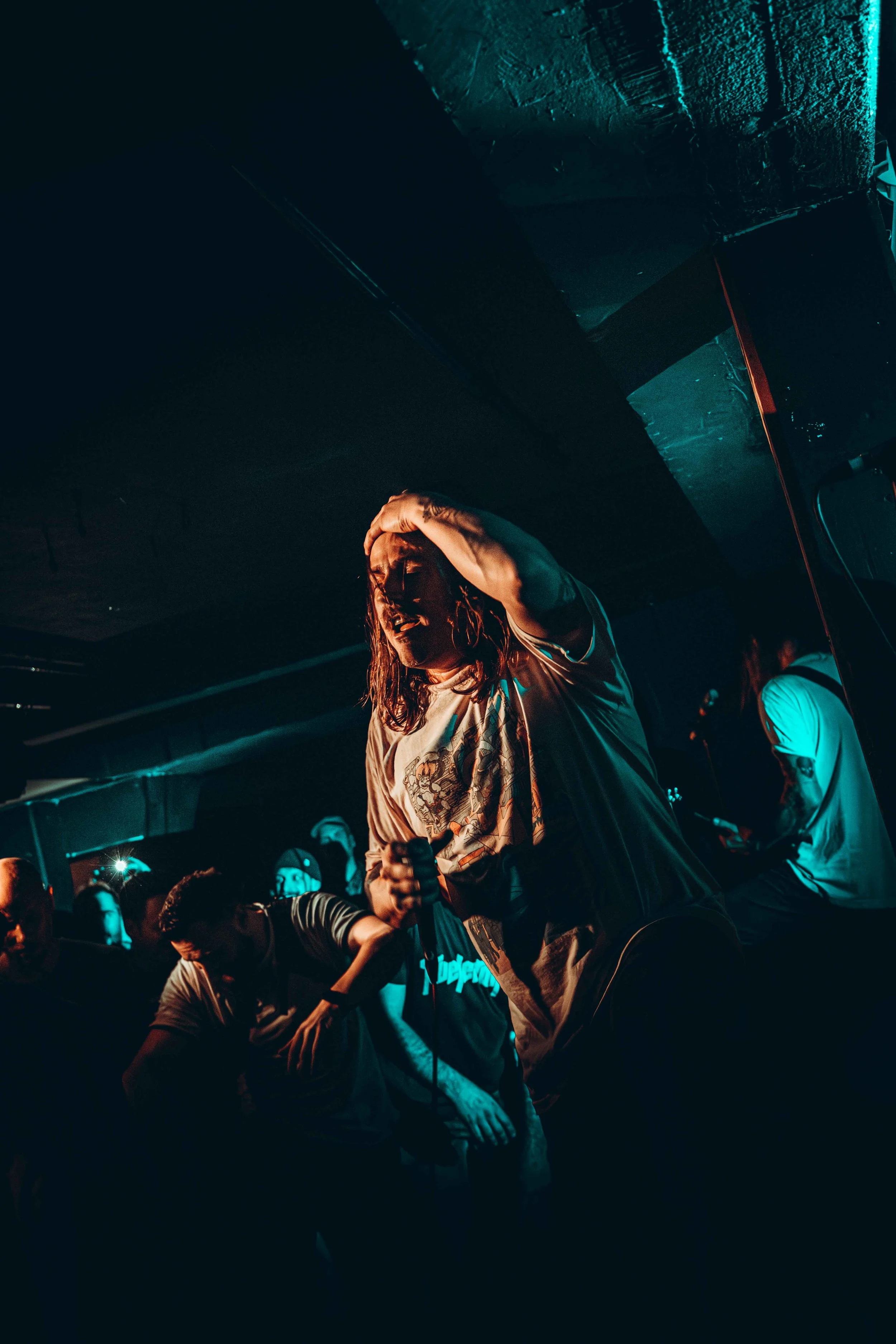 A male performer with long hair holding a microphone on stage during a concert, with dark lighting and an audience in the background.
