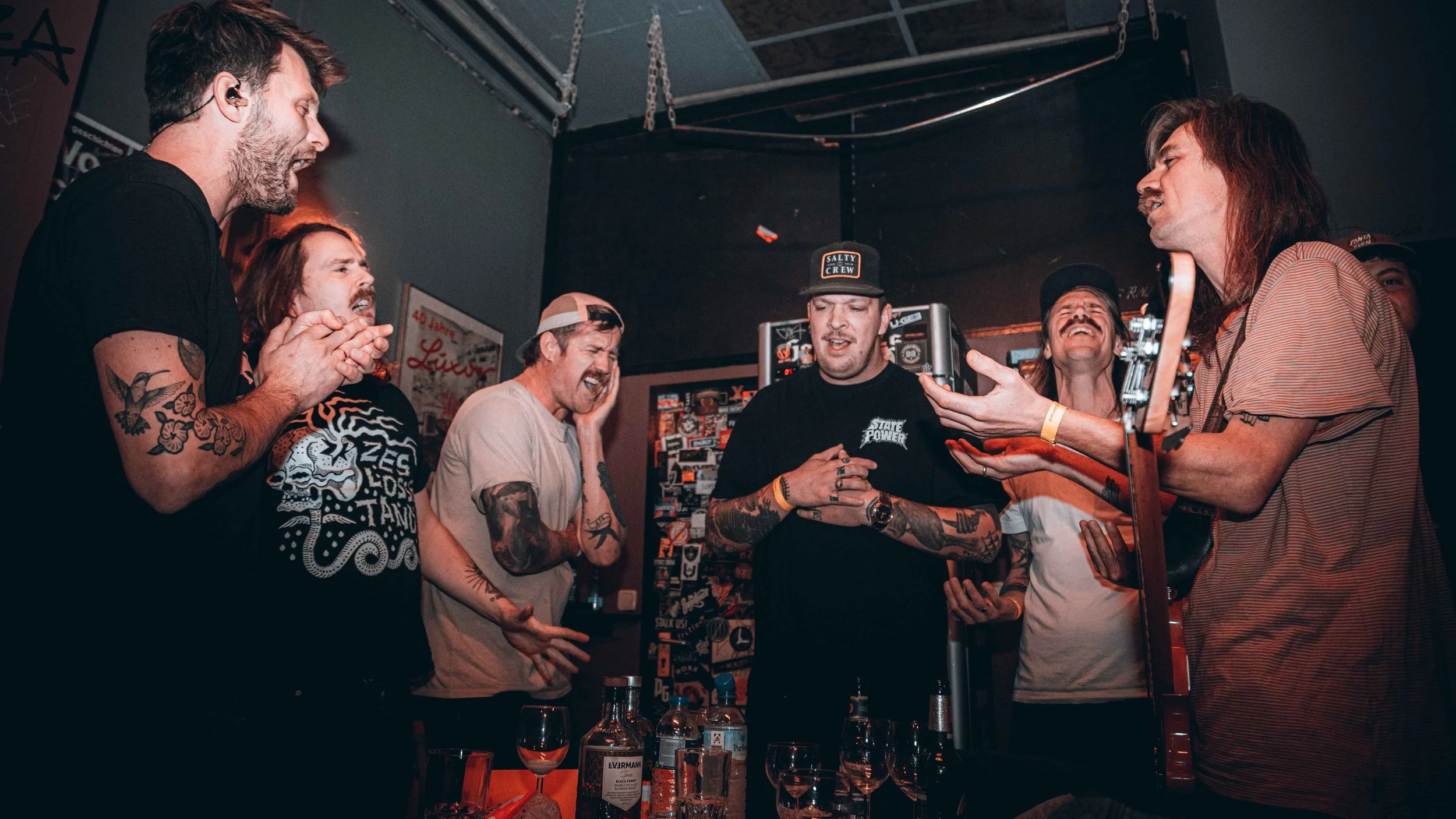 Group of men with tattoos singing and celebrating at a bar, with one man holding a guitar.