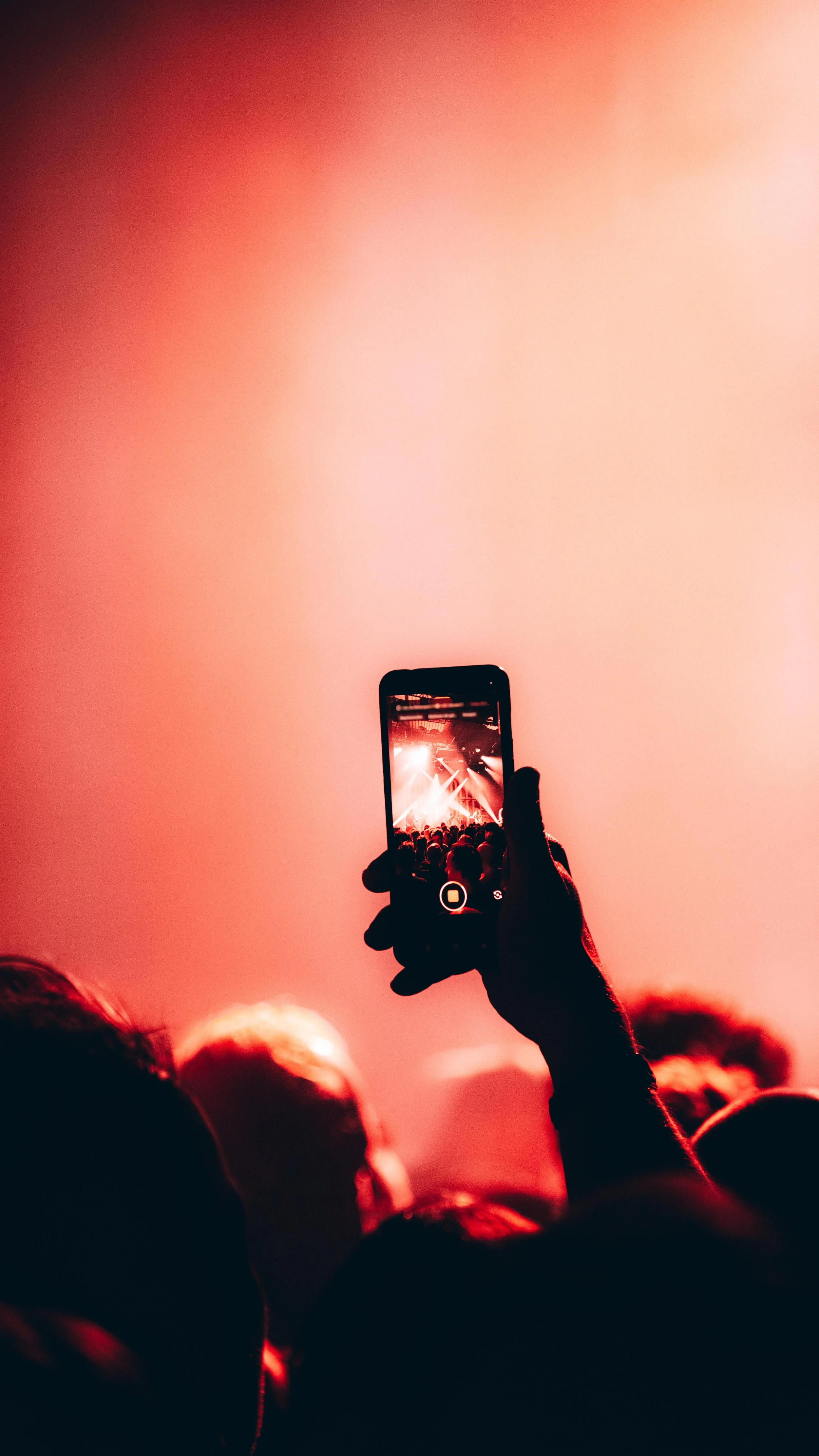 Concert audience with a person recording the event on a smartphone, with stage lights and crowd visible on the phone screen.
