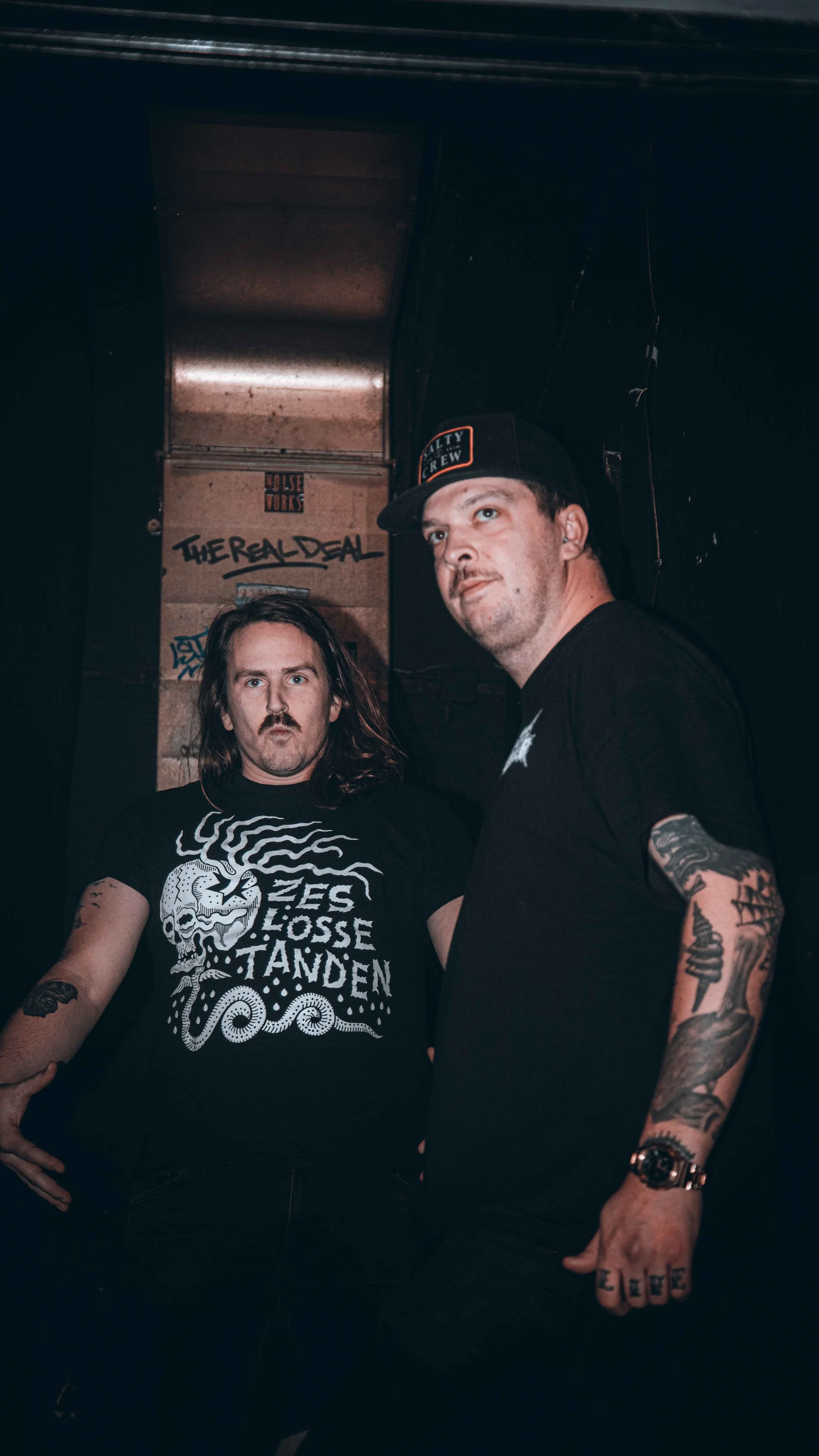 Two men with tattoos and long hair standing together inside a dark room with graffiti on the wall behind them. One man is wearing a black T-shirt with a skull and octopus design and the words "LES LOSE TAVDEN." The other man is wearing a black cap an